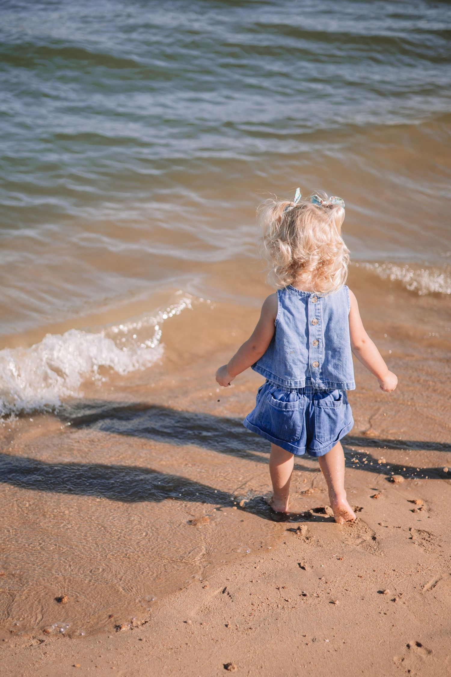 Small child in blue romper plays on sandy beach near ocean waves on sunny day.