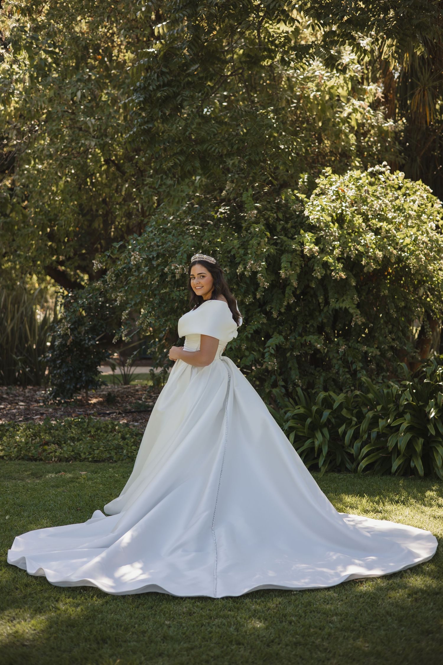 A bride in a flowing white ballgown sits gracefully on lush green grass in a sunlit garden setting.
