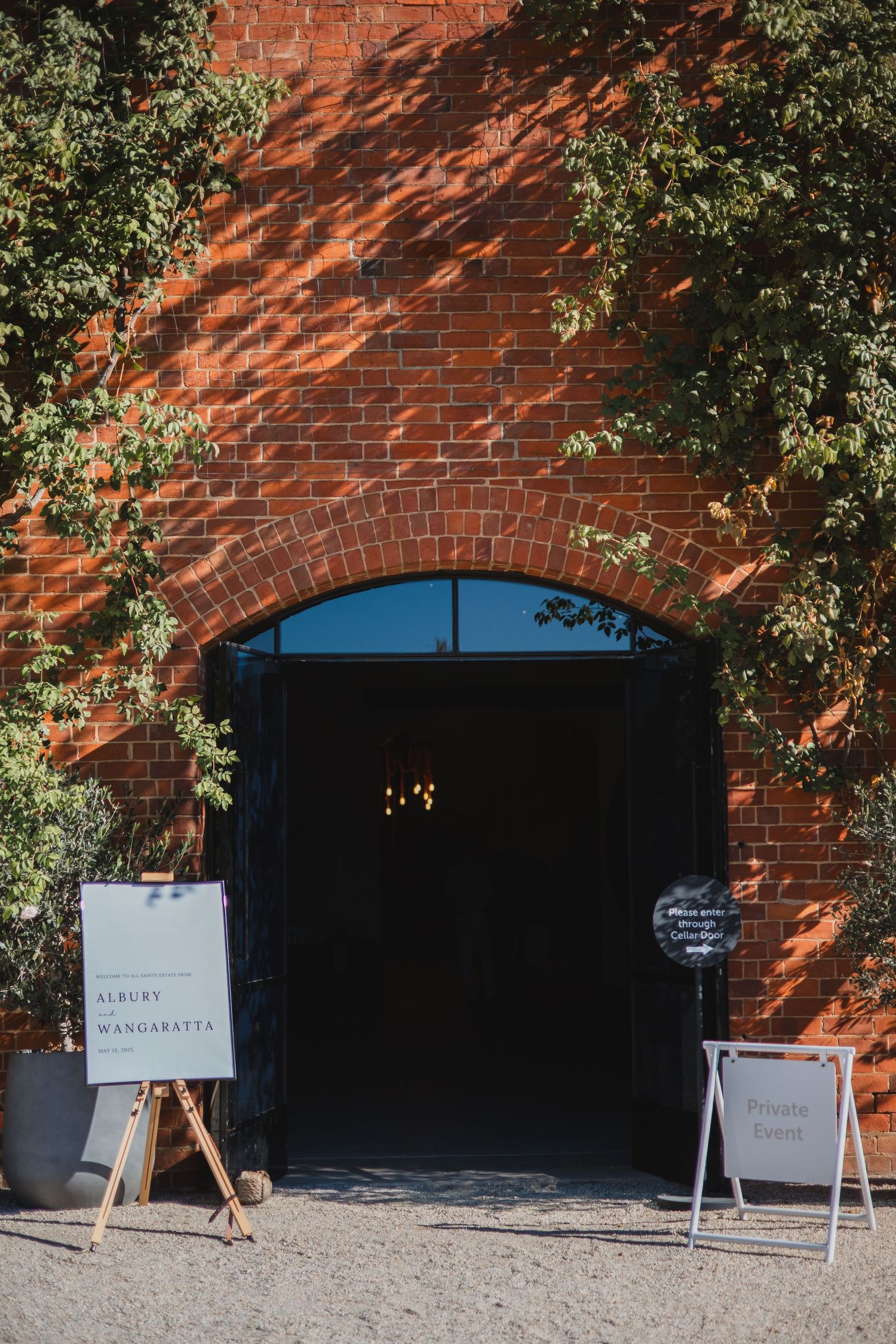 A rustic brick archway entrance with black doors and menu signboards stands framed by green foliage at an event.