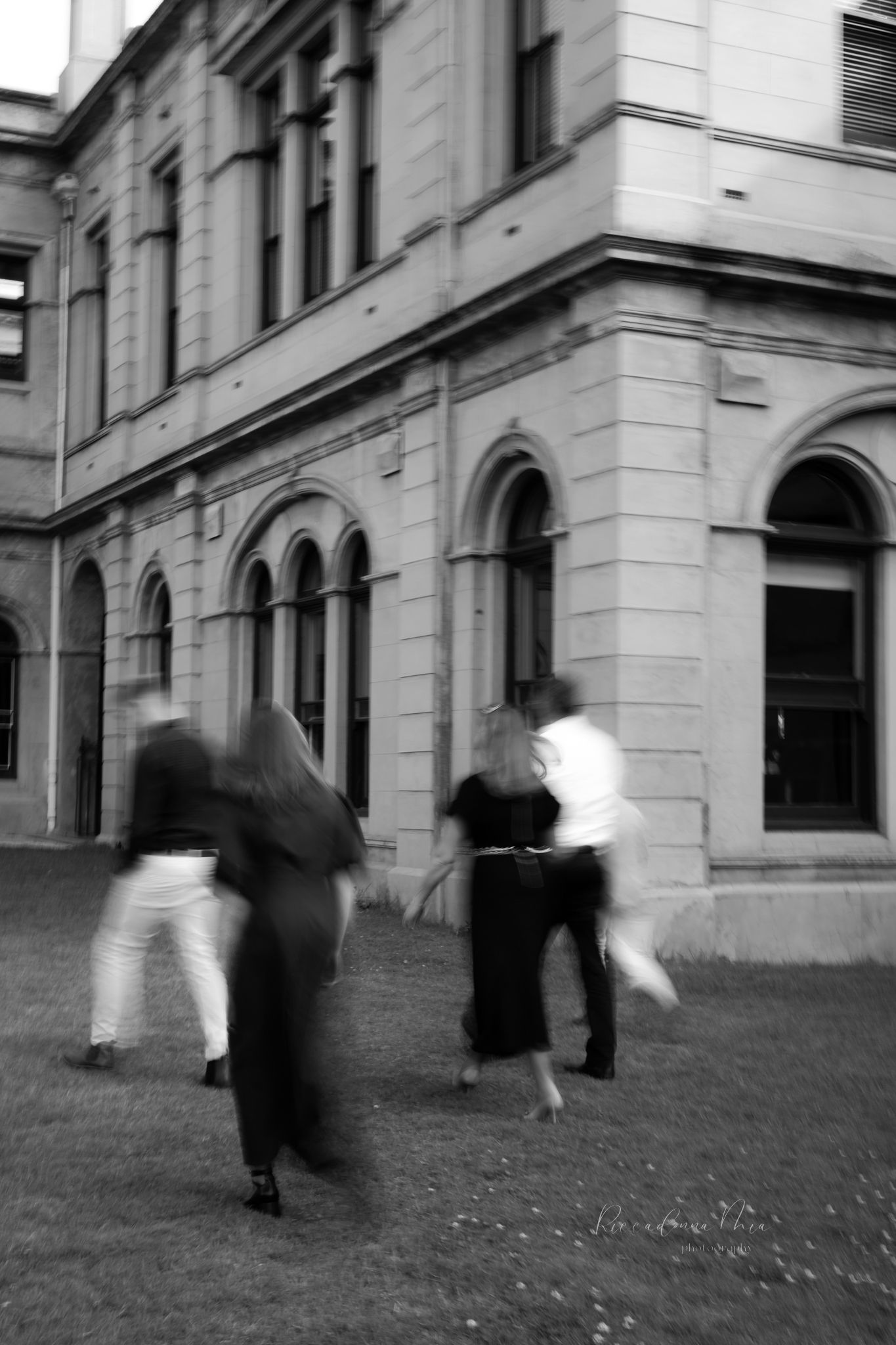 Motion-blurred figures walk past a historic stone building in black and white, creating a ghostly urban scene.