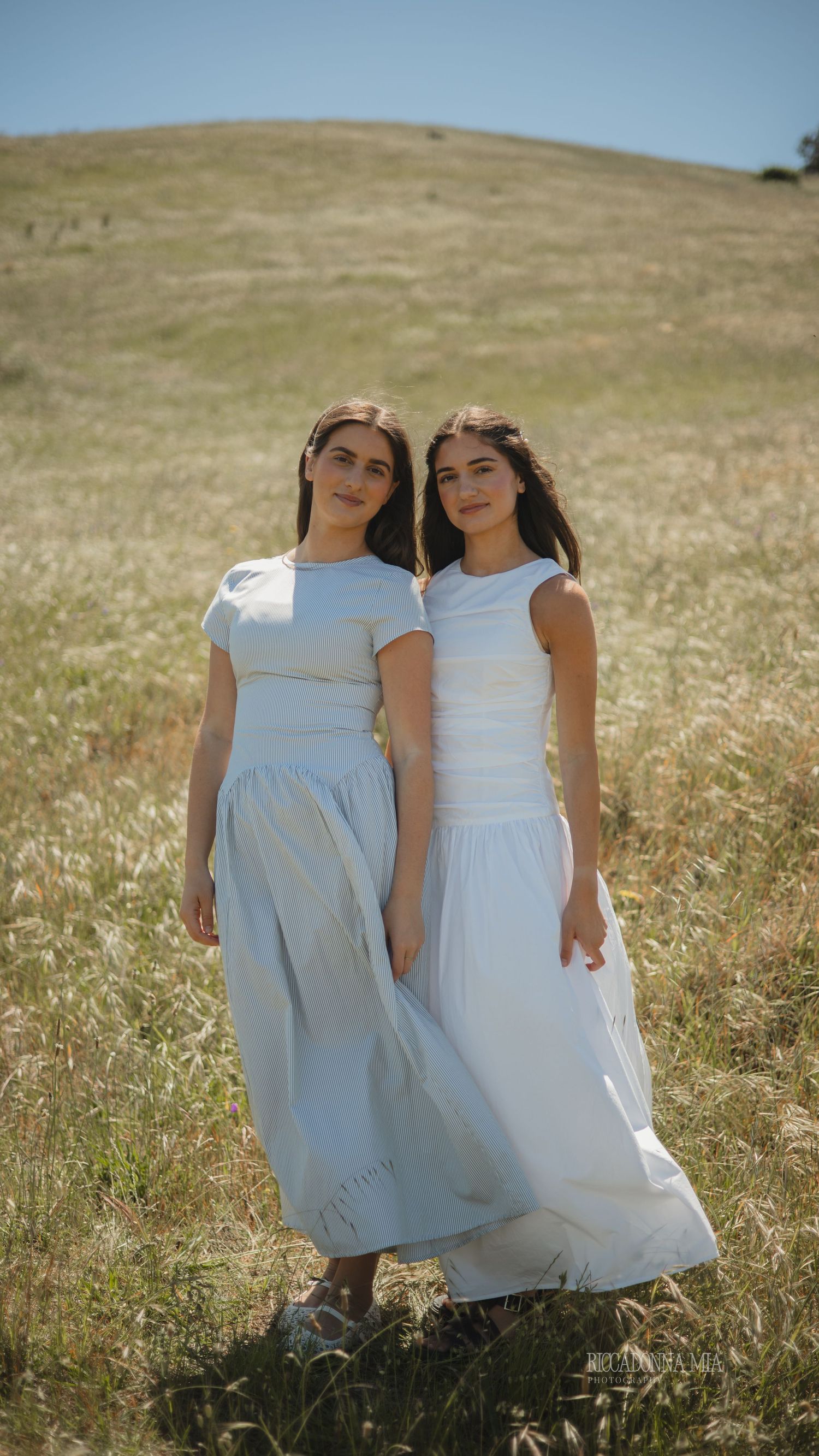 Two people in white dresses stand together in a sunlit meadow on a summer day.