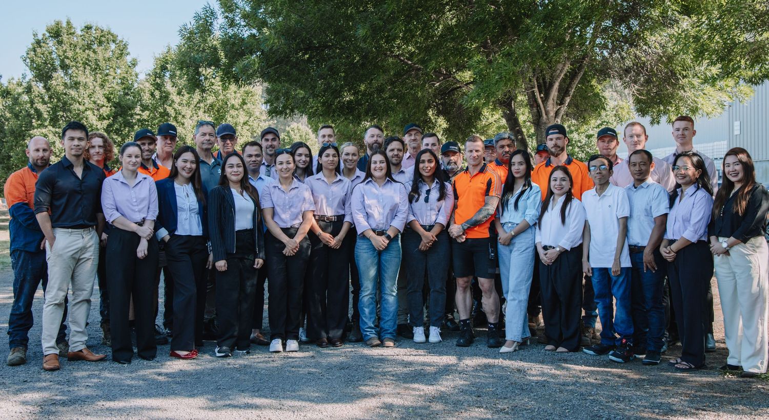 Large group of people dressed in business casual attire gathered outdoors under trees for a company photo.