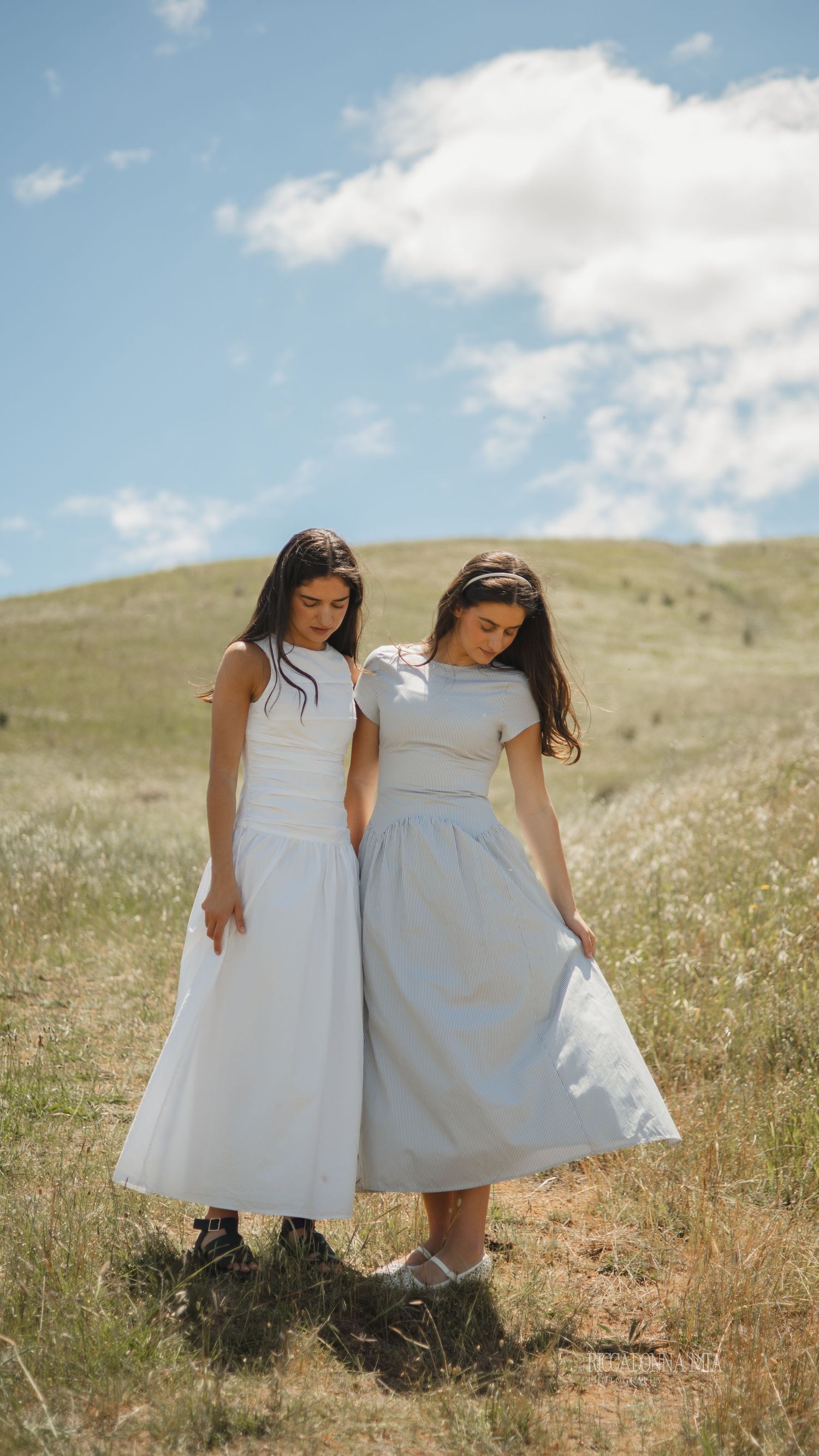 Two young girls in white dresses and boots stand together in a sunny meadow with wildflowers and rolling hills.