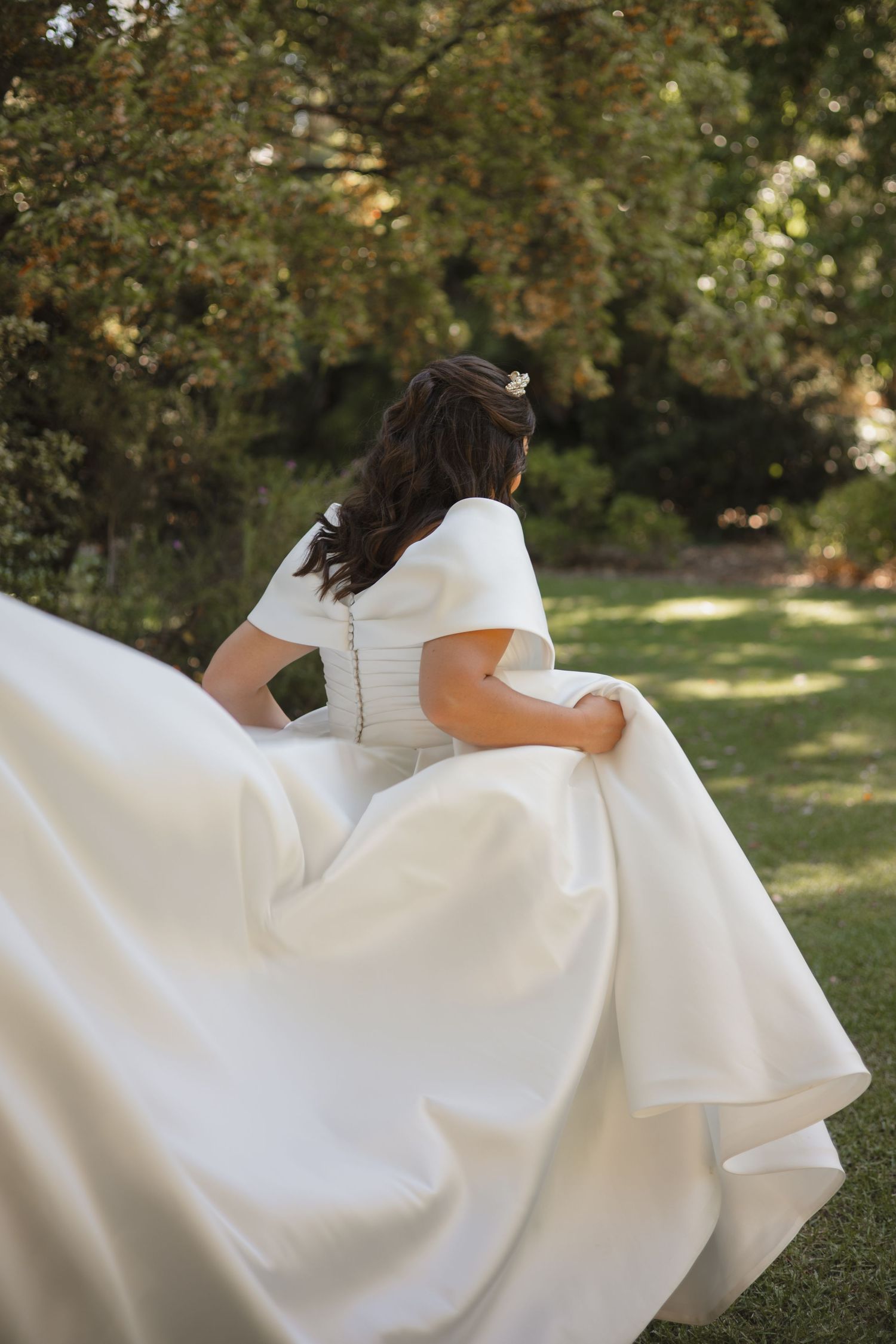 Back view of a flowing white wedding dress with short sleeves in a sunlit garden setting.