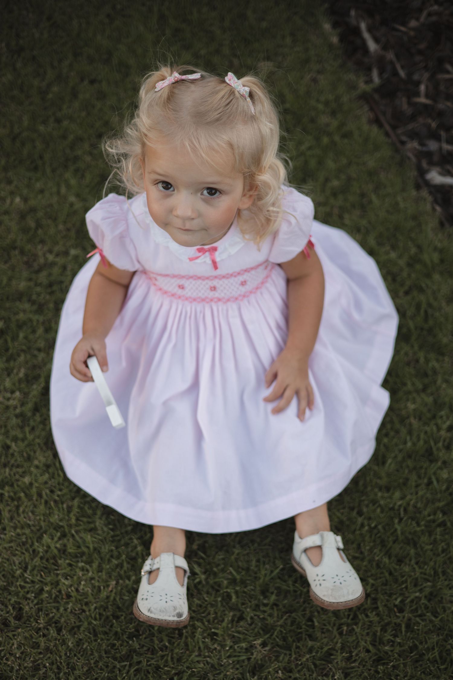 Young child in white smocked dress with pink details and white sandals poses on grass.