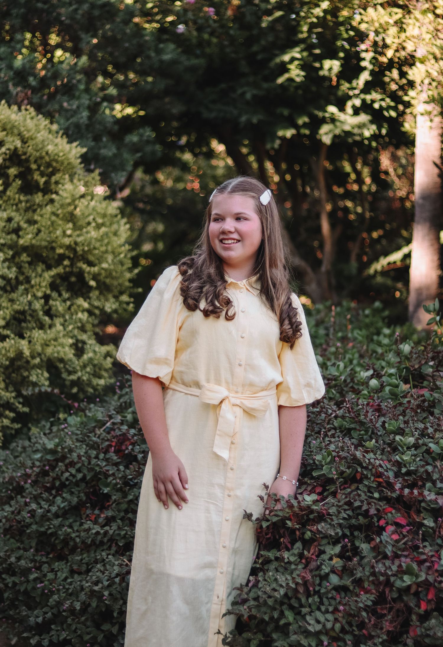 Person in a vintage-inspired pale yellow dress with puff sleeves and waist tie poses outdoors in a garden setting.