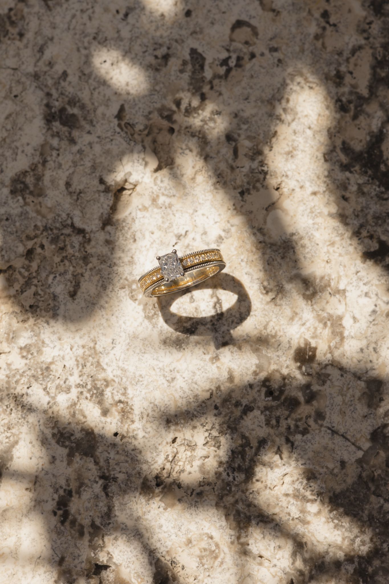 A diamond engagement ring casts dramatic shadows on a textured stone or concrete surface in natural lighting.