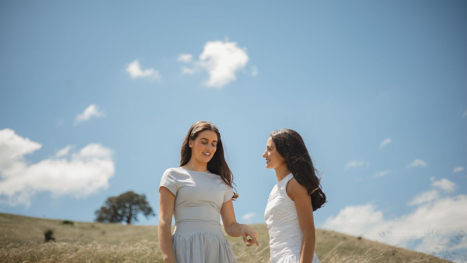 Two women in white dresses stand together on a grassy hilltop against a bright blue sky with puffy clouds.