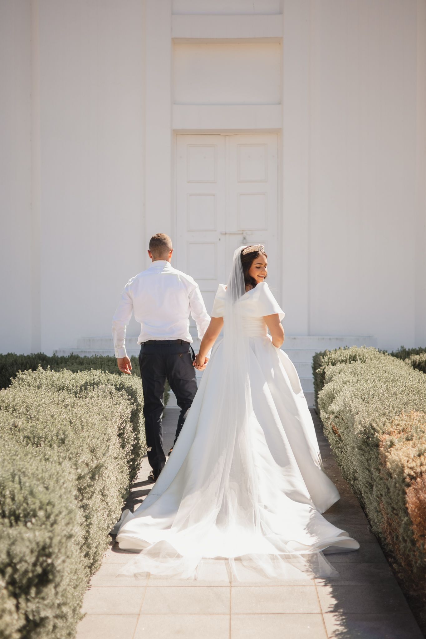 Newlywed couple walking along garden path with bride in flowing white gown and train on sunny day.