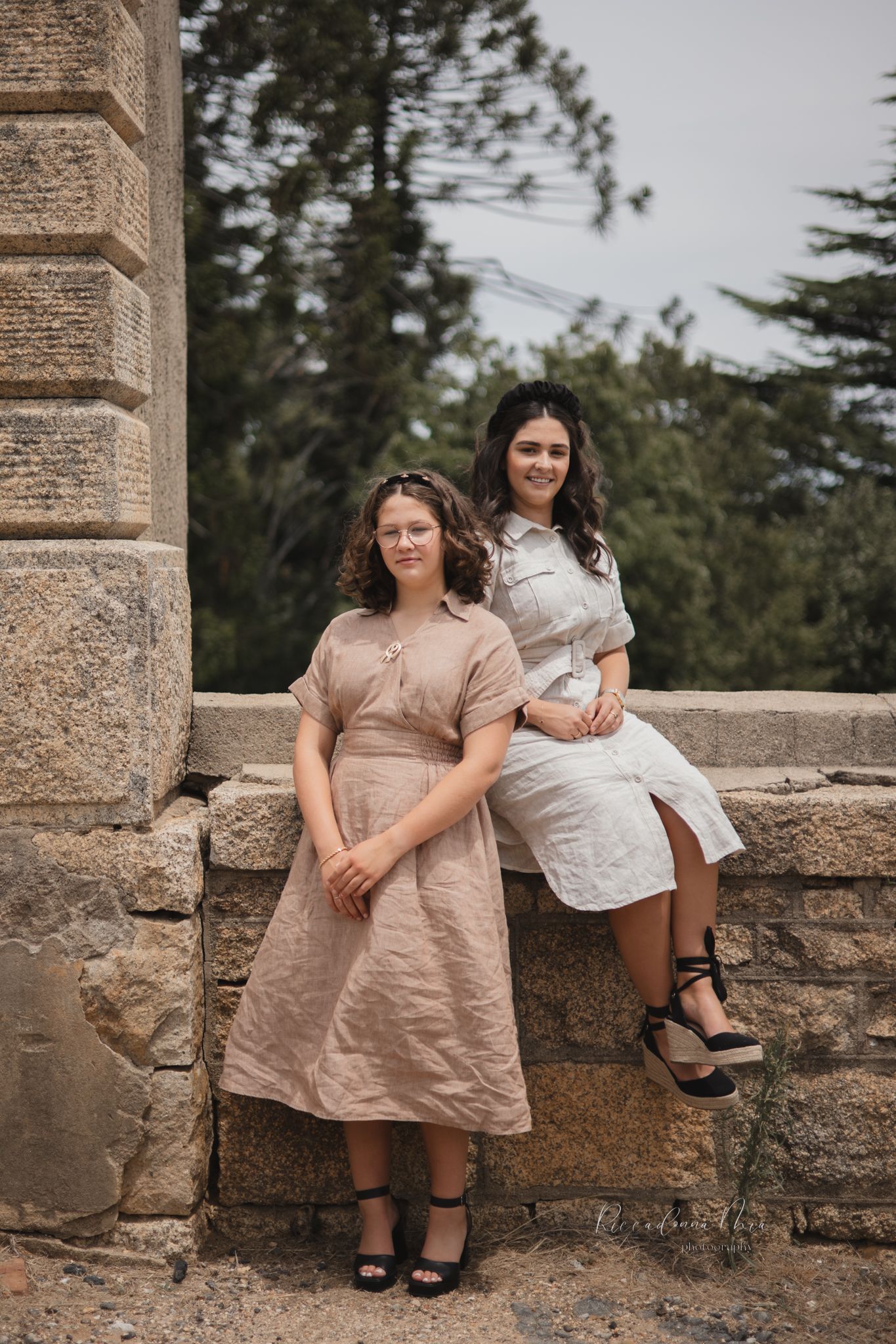 Two women in vintage-style dresses pose by a rustic stone wall amid trees and greenery.