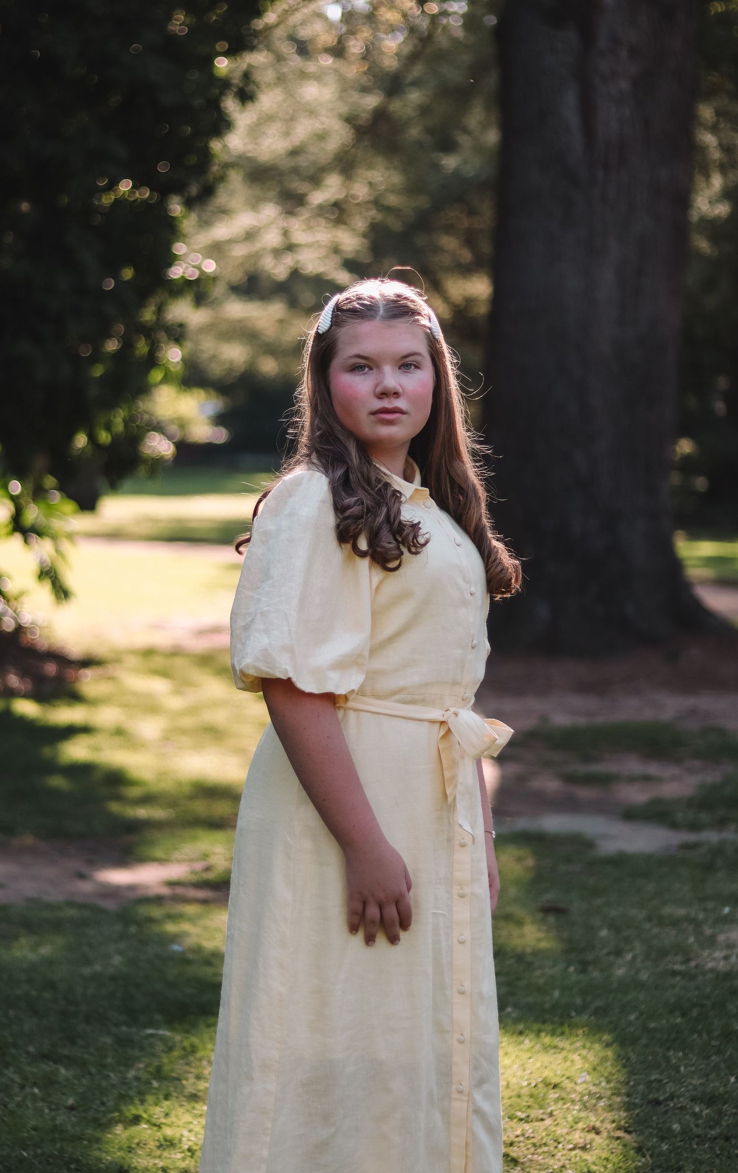 Person in cream-colored vintage dress standing in sunlit garden with trees in background.