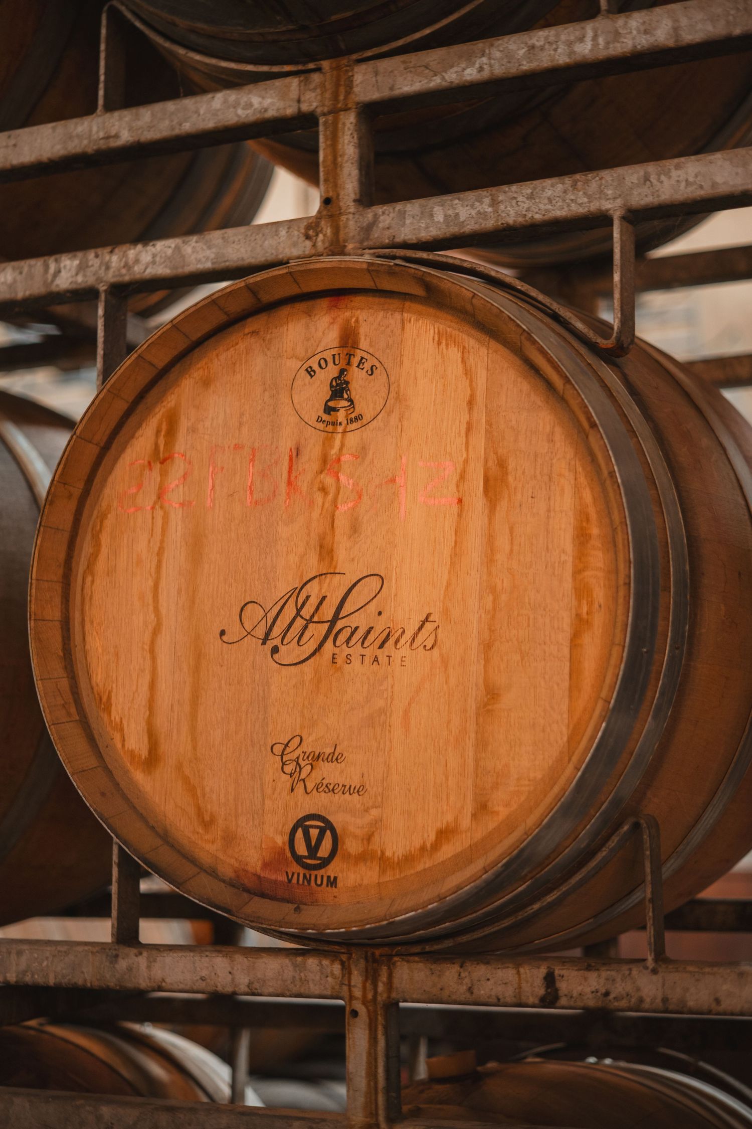 Wooden wine barrel with All Saints branding stacked in a winery cellar with metal rack supports.