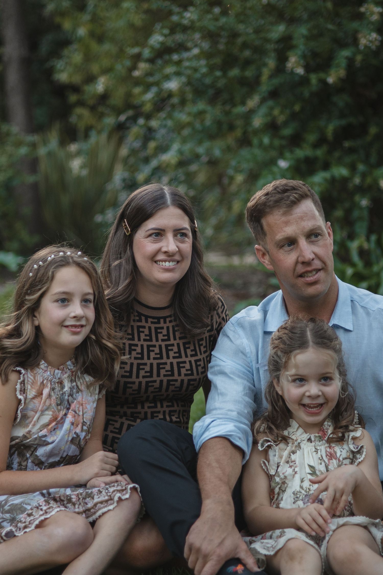 A family of four sits together outdoors on a sunny day against a green foliage backdrop.