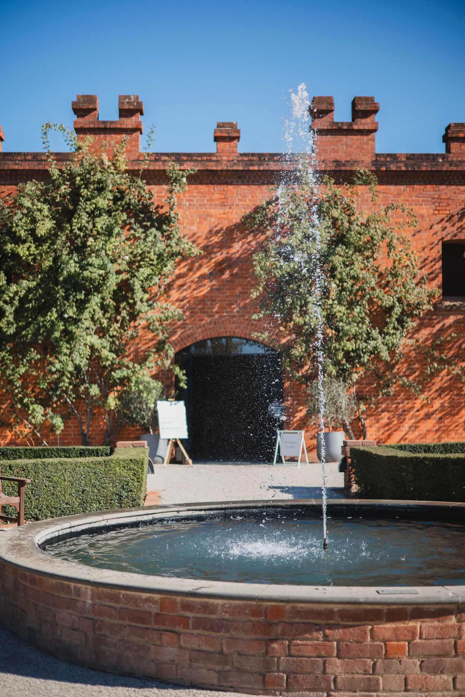 A brick-walled courtyard features a circular fountain with water spray and decorative hedge landscaping around an archway entrance.