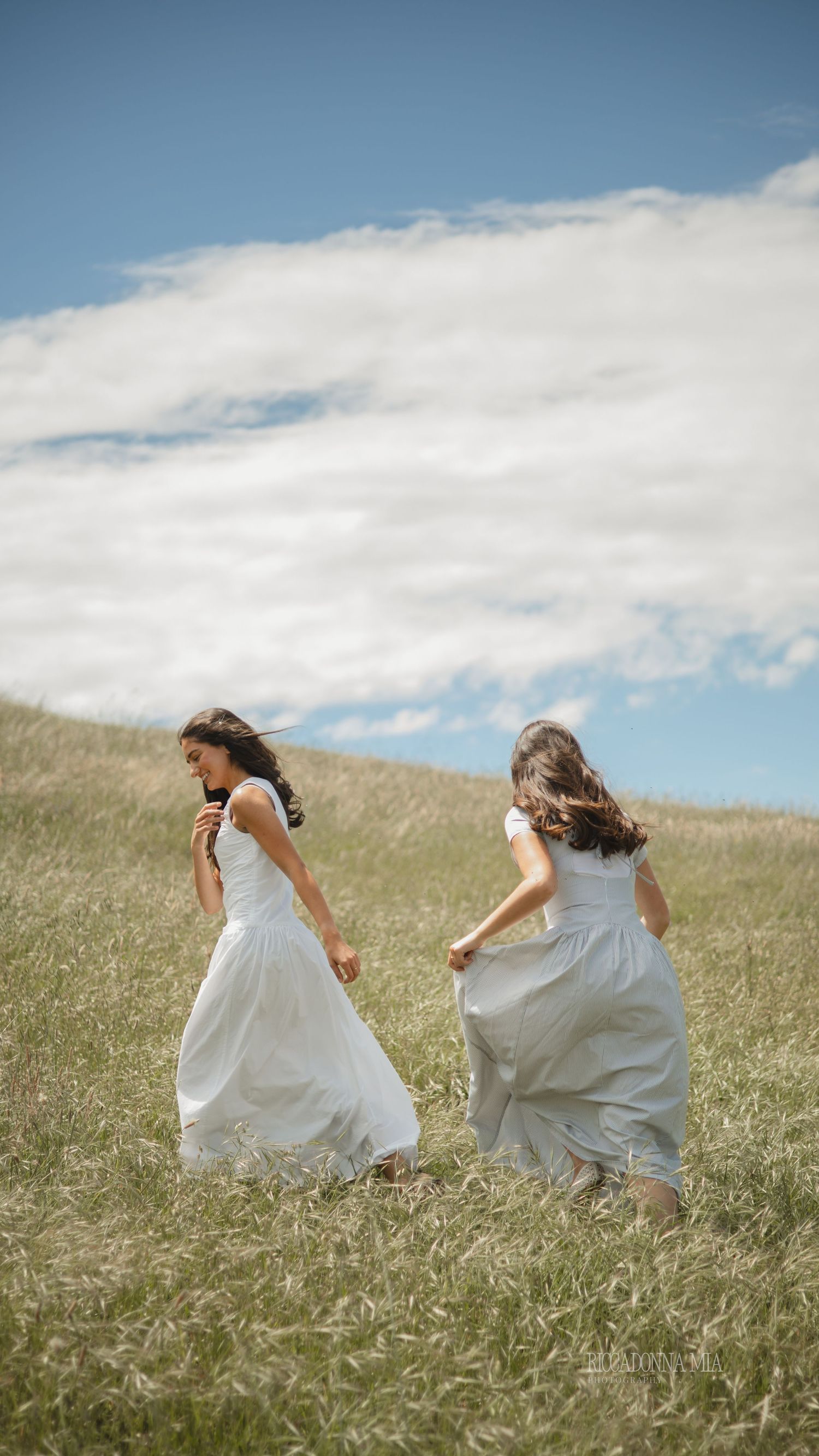 Two people in white dresses run playfully through a grassy meadow under a cloudy summer sky.