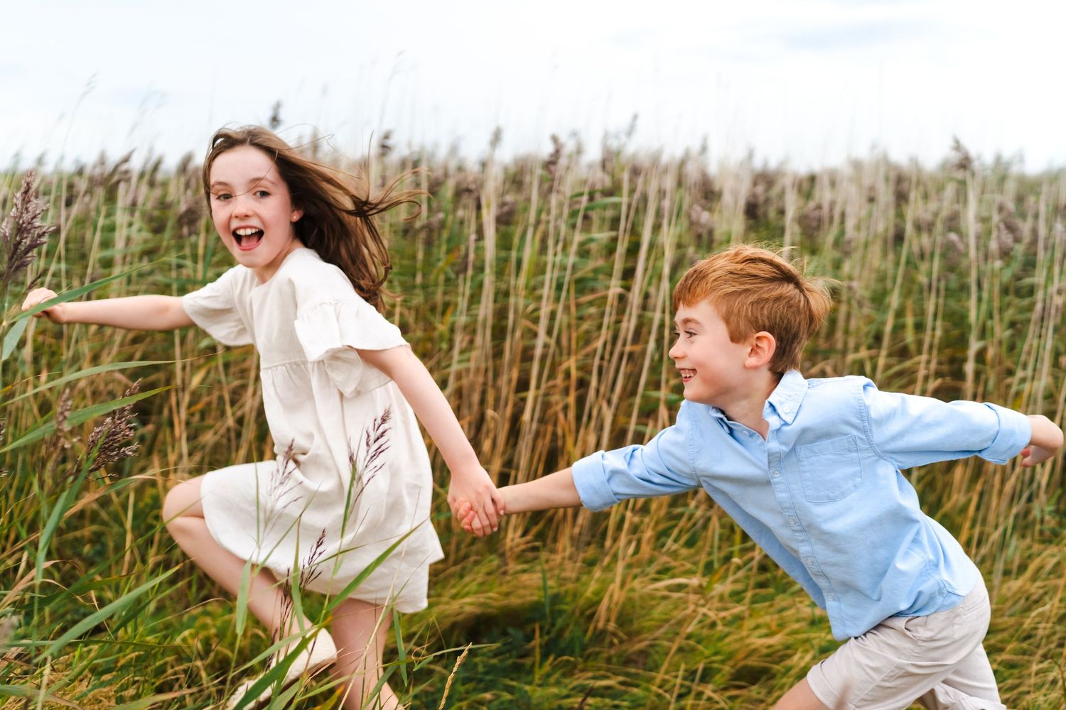 Playful family running and jumping through tall grass on a sunny beach path.  Family photographer Dublin Ireland