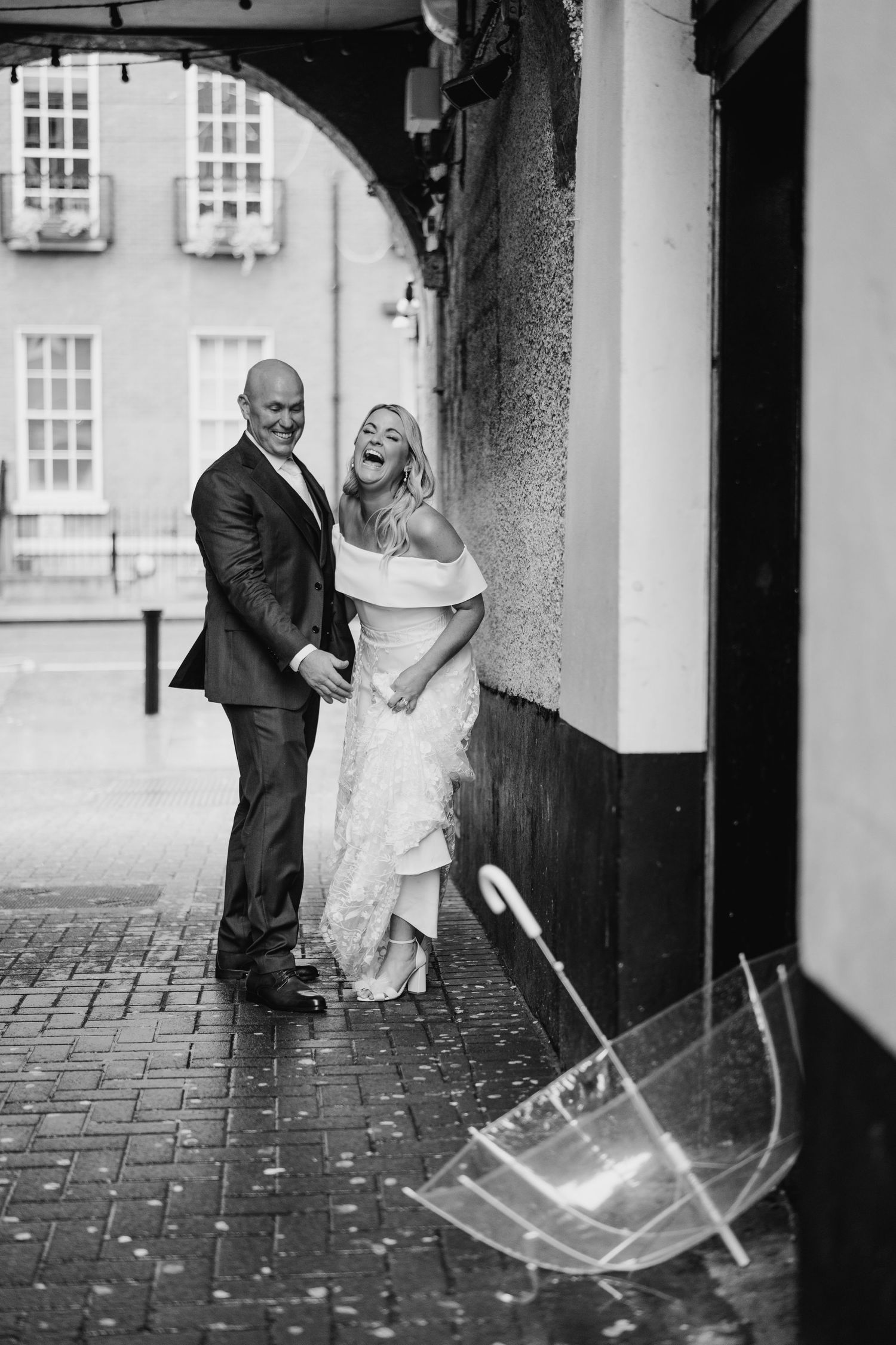 A couple poses with clear umbrellas on a rainy cobblestone walkway outside a historic building. The Dean Dublin Wedding Photographer