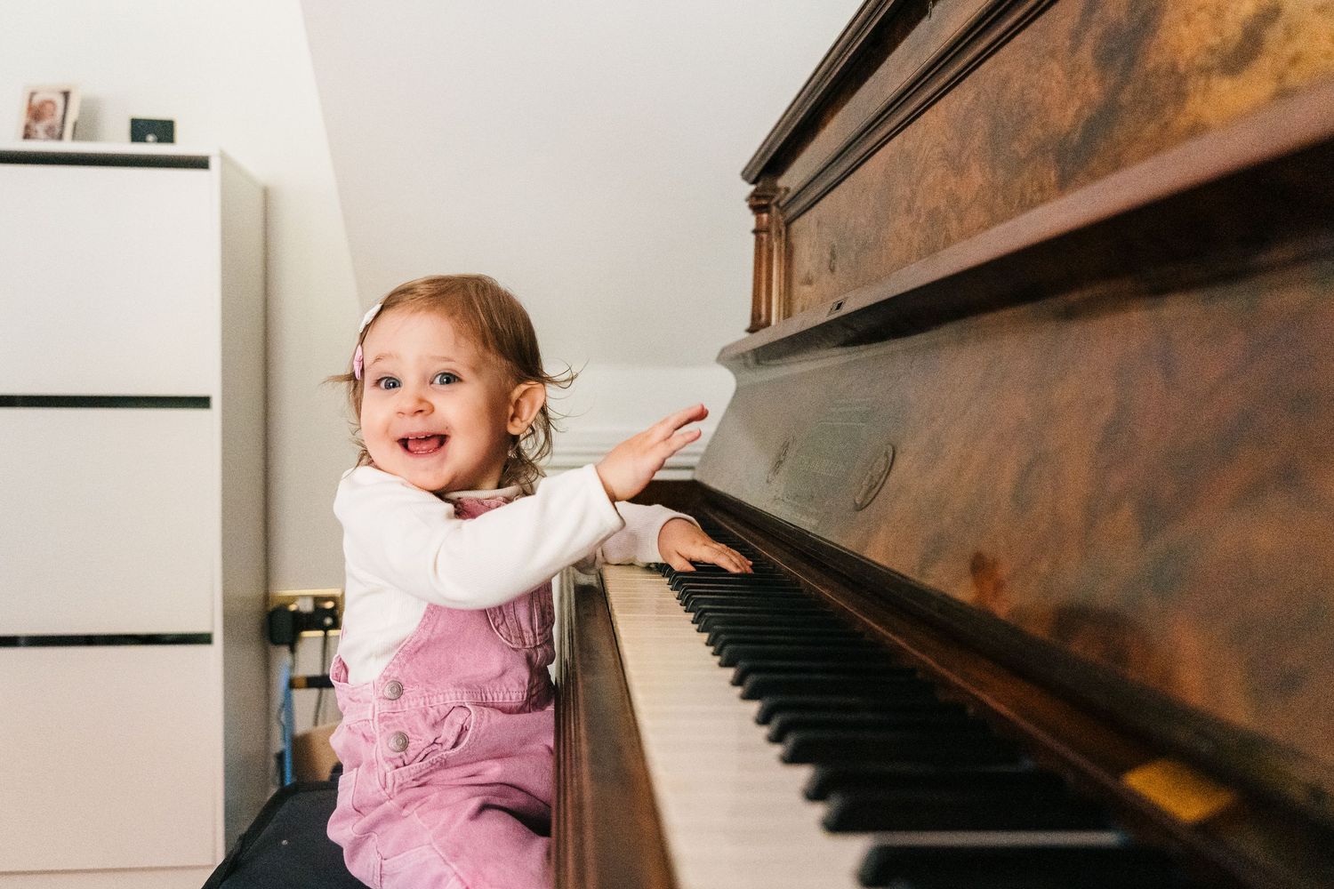 A small child in pink overalls reaches up to play an upright wooden piano in a home setting. family PHOTOGRAPHER Dublin