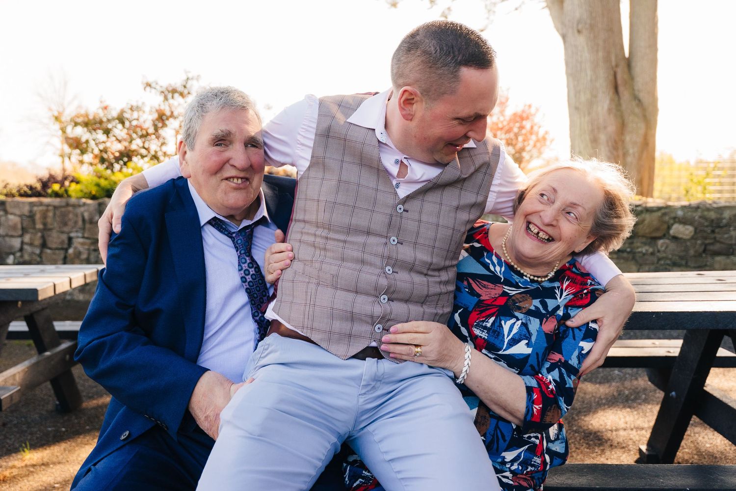 Three people sit together on a wooden bench at sunset sharing joyful moments in a park. family PHOTOGRAPHER Dublin