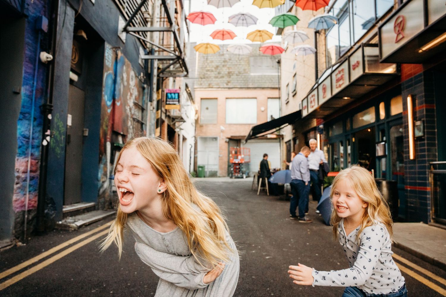 Children run joyfully beneath colorful umbrellas hanging overhead on a narrow urban street in Dublin. family PHOTOGRAPHER Dublin