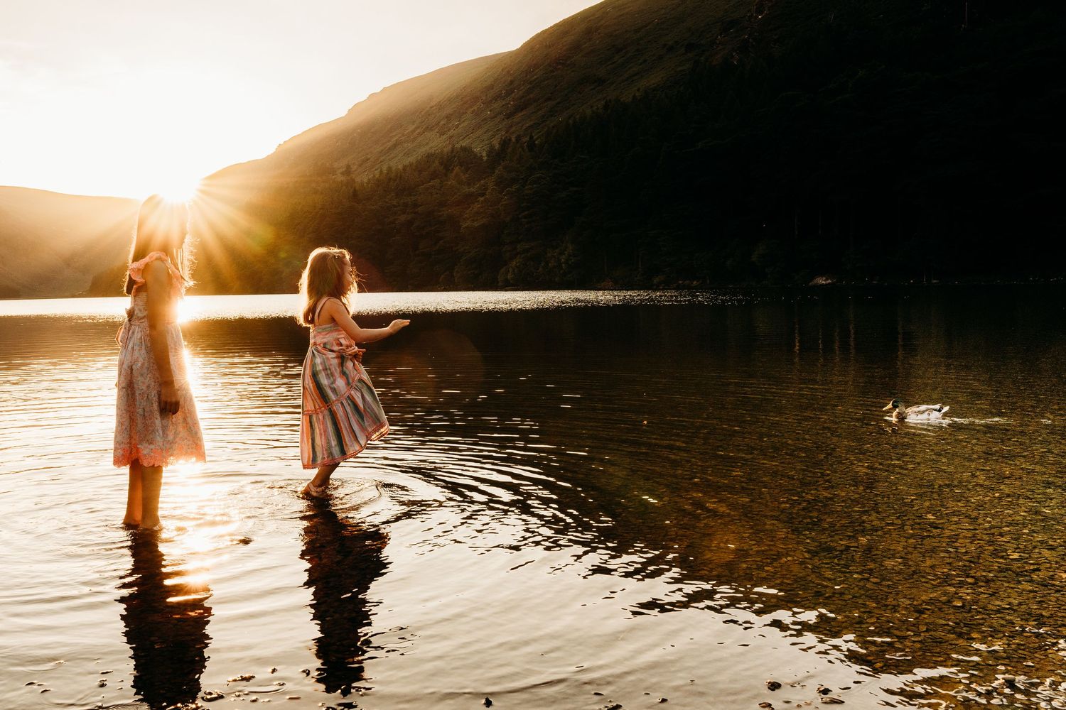 Two people wade through calm lake water at sunset with sunlight creating a golden silhouette effect. family PHOTOGRAPHER Dublin