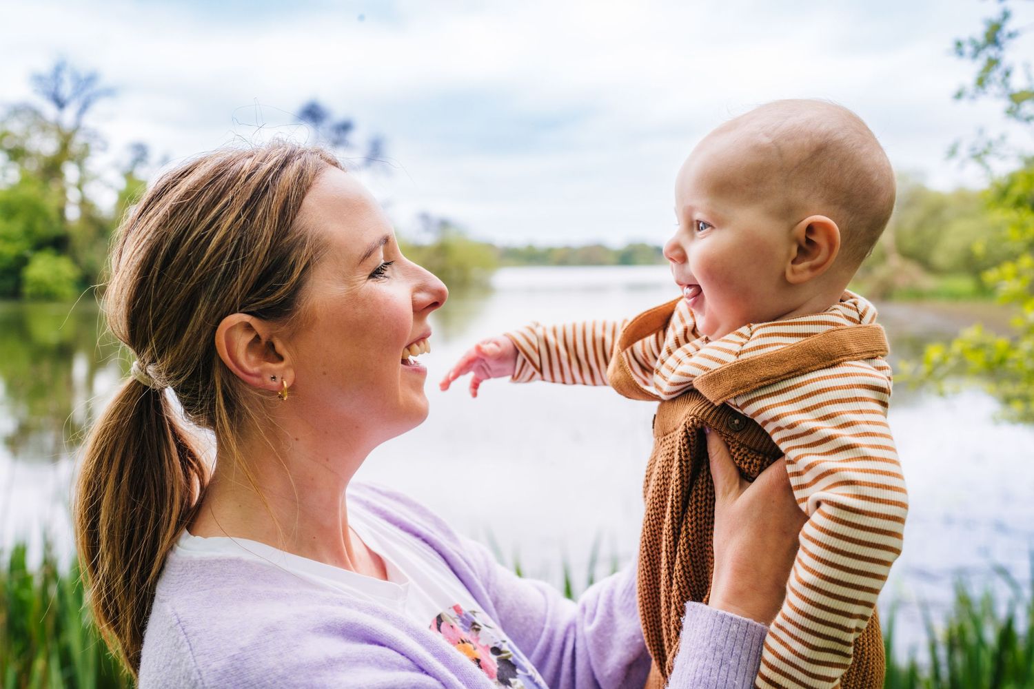 A person lifts and spins a laughing child in front of a lake on a sunny day while sharing sweet moments together. family PHOTOGRAPHER Dublin