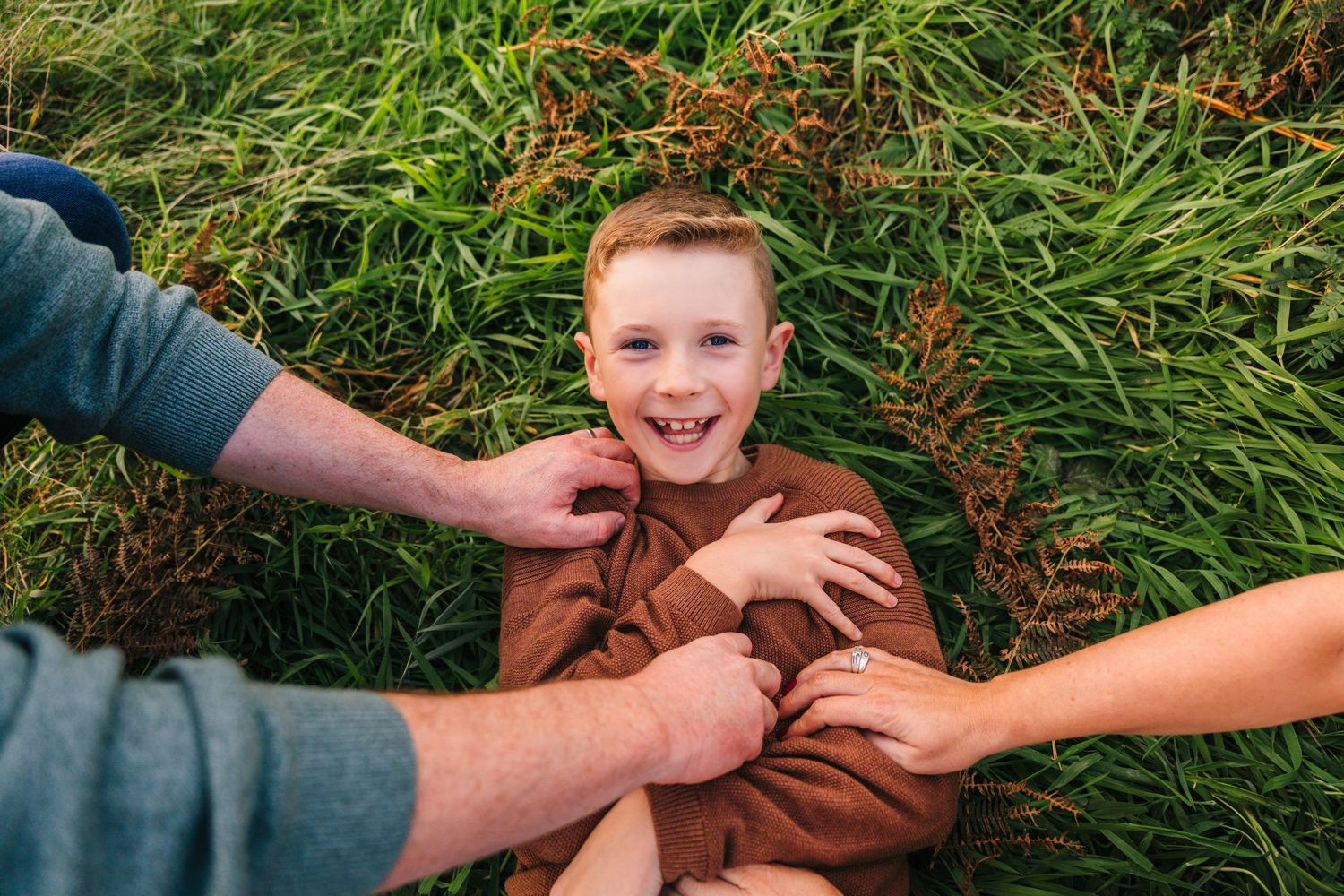 Someone laying in grass is being playfully tickled by two hands reaching from opposite sides. family PHOTOGRAPHER Dublin