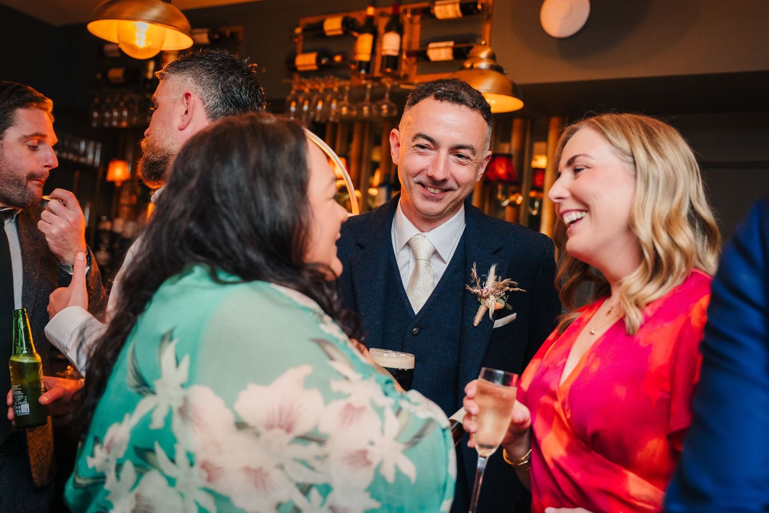 A candid moment of wedding guests laughing and socializing at a bar reception.