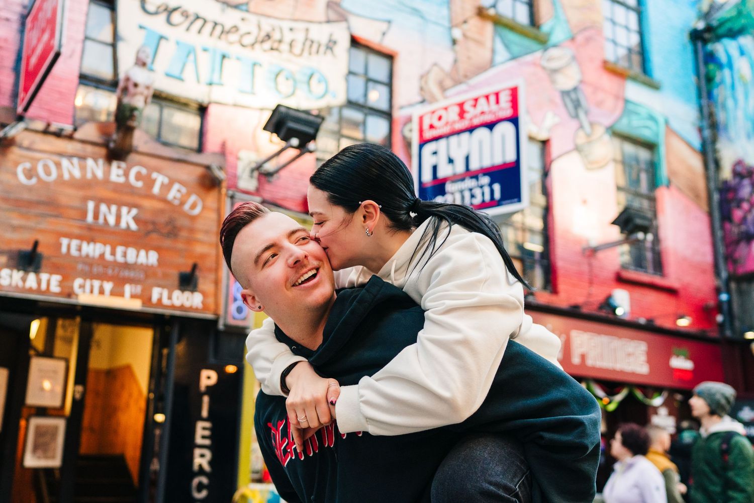 Two people share an intimate moment on a busy street with colorful storefronts and neon signs behind them. PHOTOGRAPHER Dublin PHOTOGRAPHER