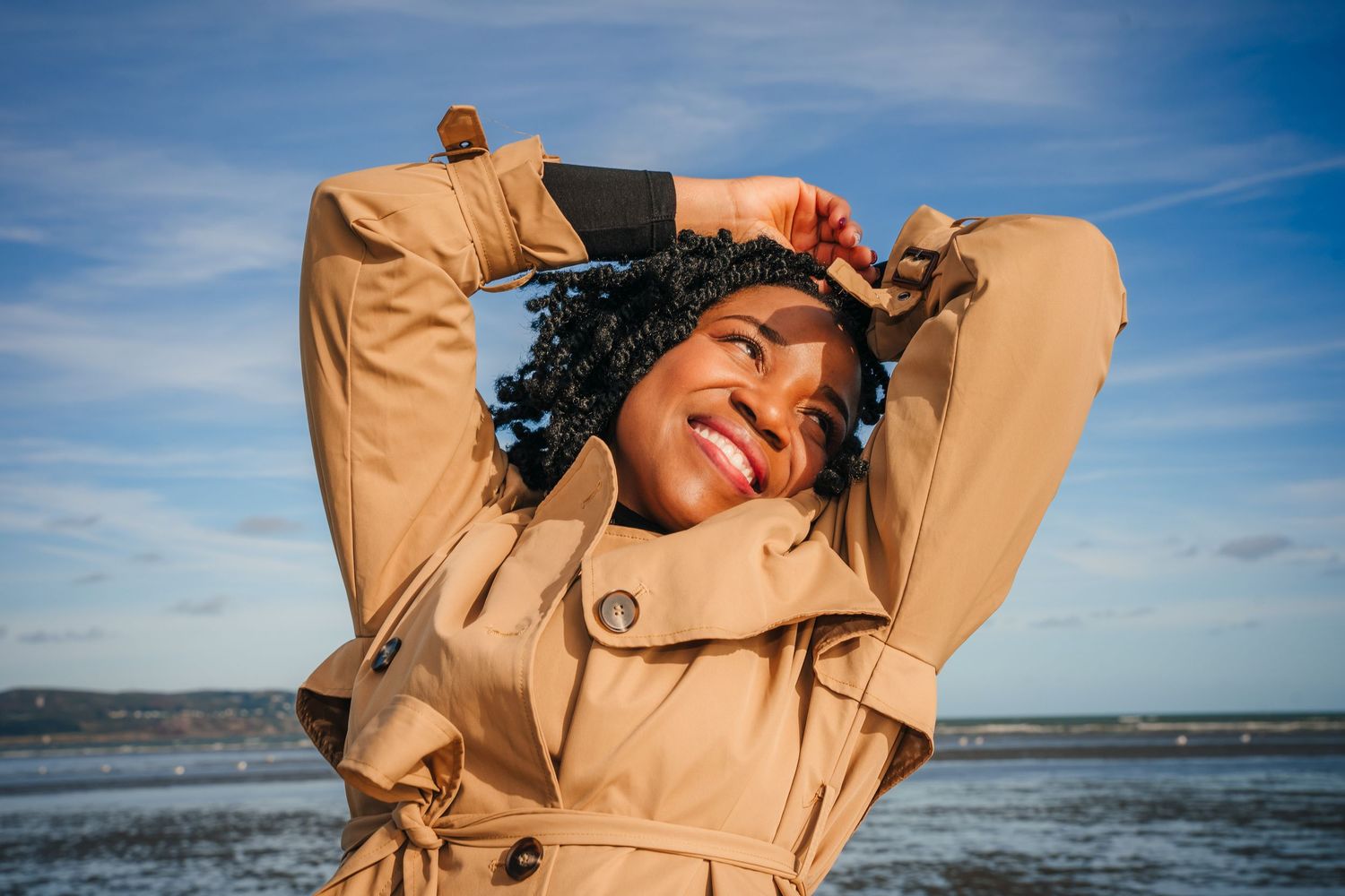 Figure in tan clothing poses expressively by the ocean on a bright sunny day. PHOTOGRAPHER Dublin ireland