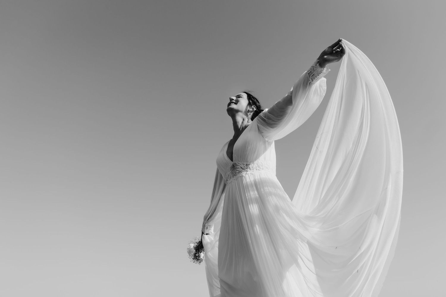 Ethereal bride in flowing white dress poses dramatically against sky as wind catches her long veil in black and white.