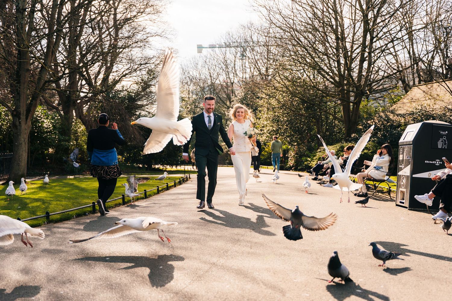 A couple walks down a sunny park path surrounded by doves and pigeons taking flight on a spring day. Wedding PHOTOGRAPHER Dublin