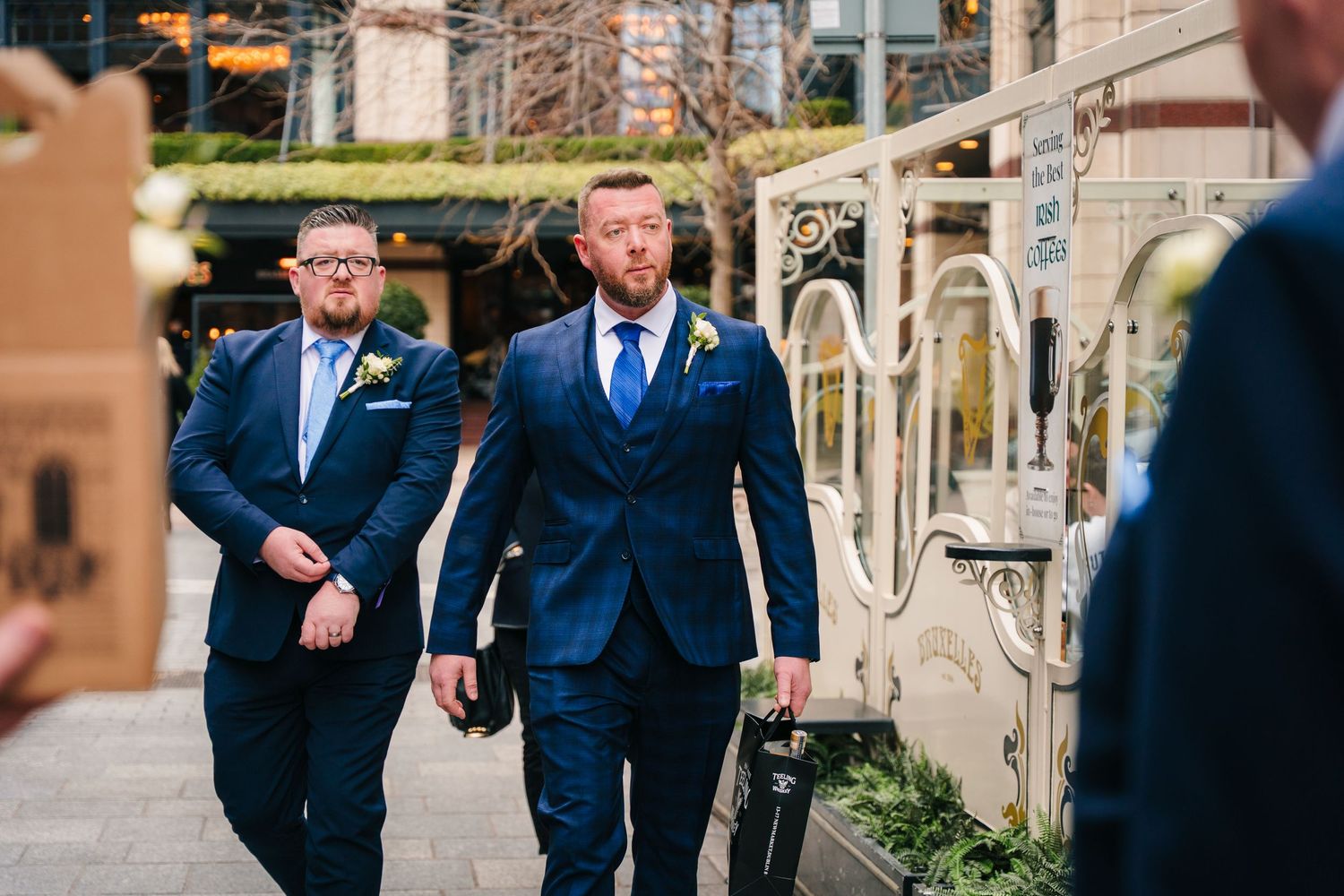Groomsmen in navy blue suits walk together and laugh while strolling along a stone pathway. Dublin wedding photogrAPher Bewleys Grafton