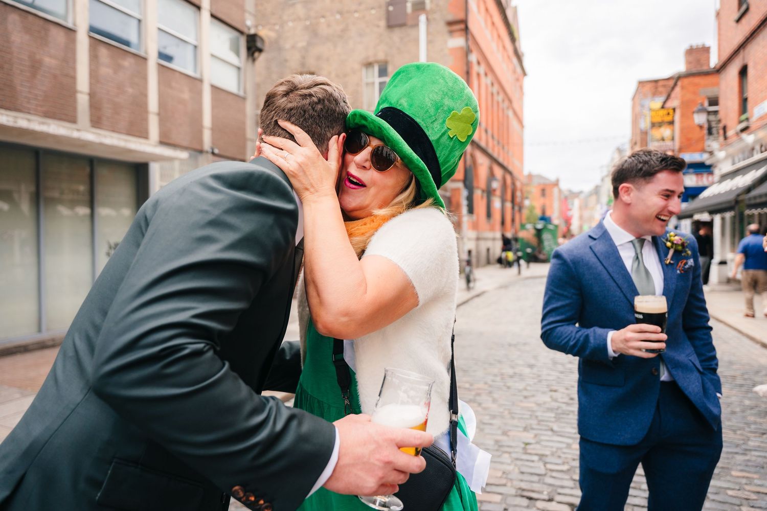 Two people embrace on a city street while wearing St Patrick's Day attire as another person stands nearby with a drink.