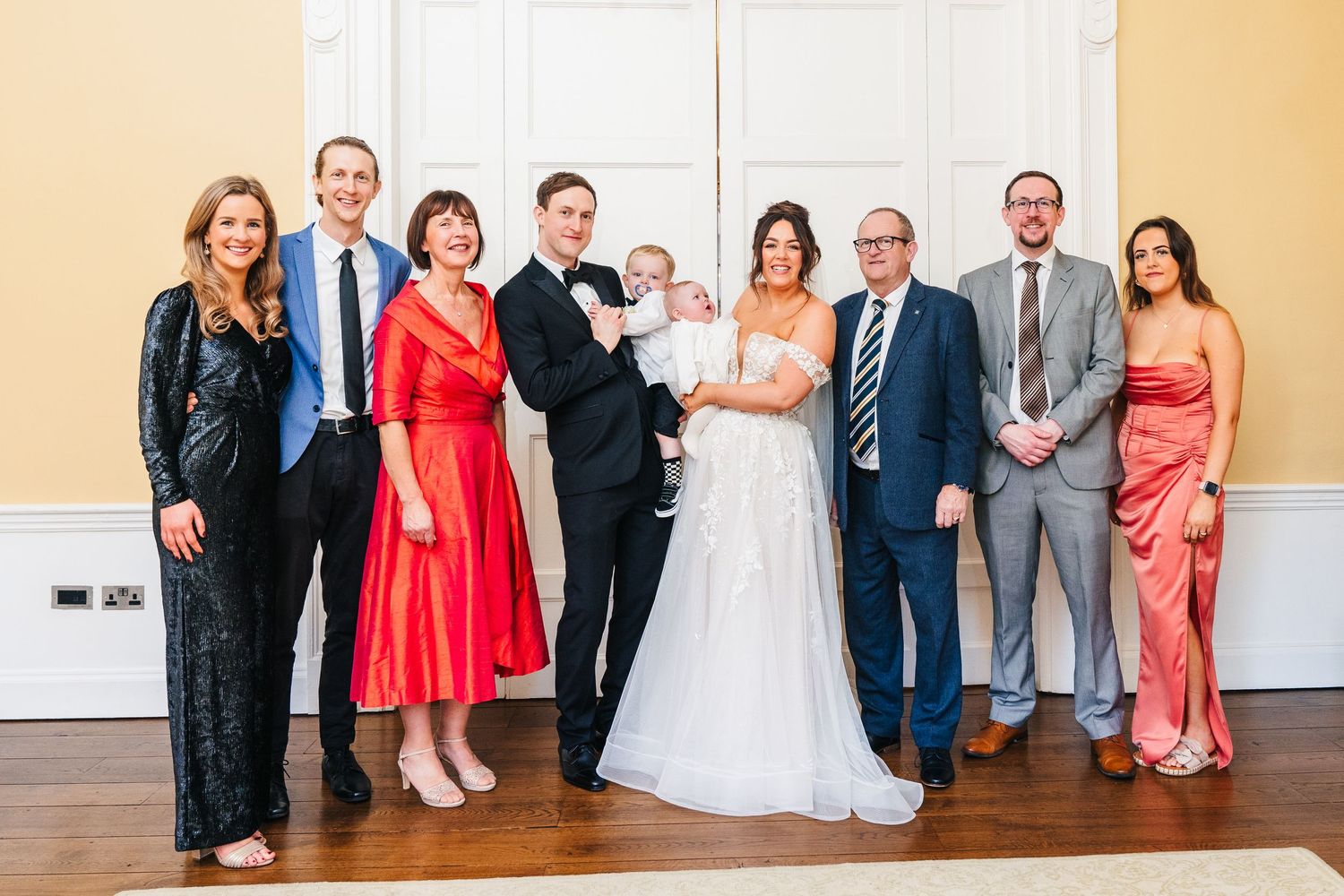 Wedding party group photo featuring coral bridesmaid dresses and formal suits in a bright room. Wedding PHOTOGRAPHER Dublin