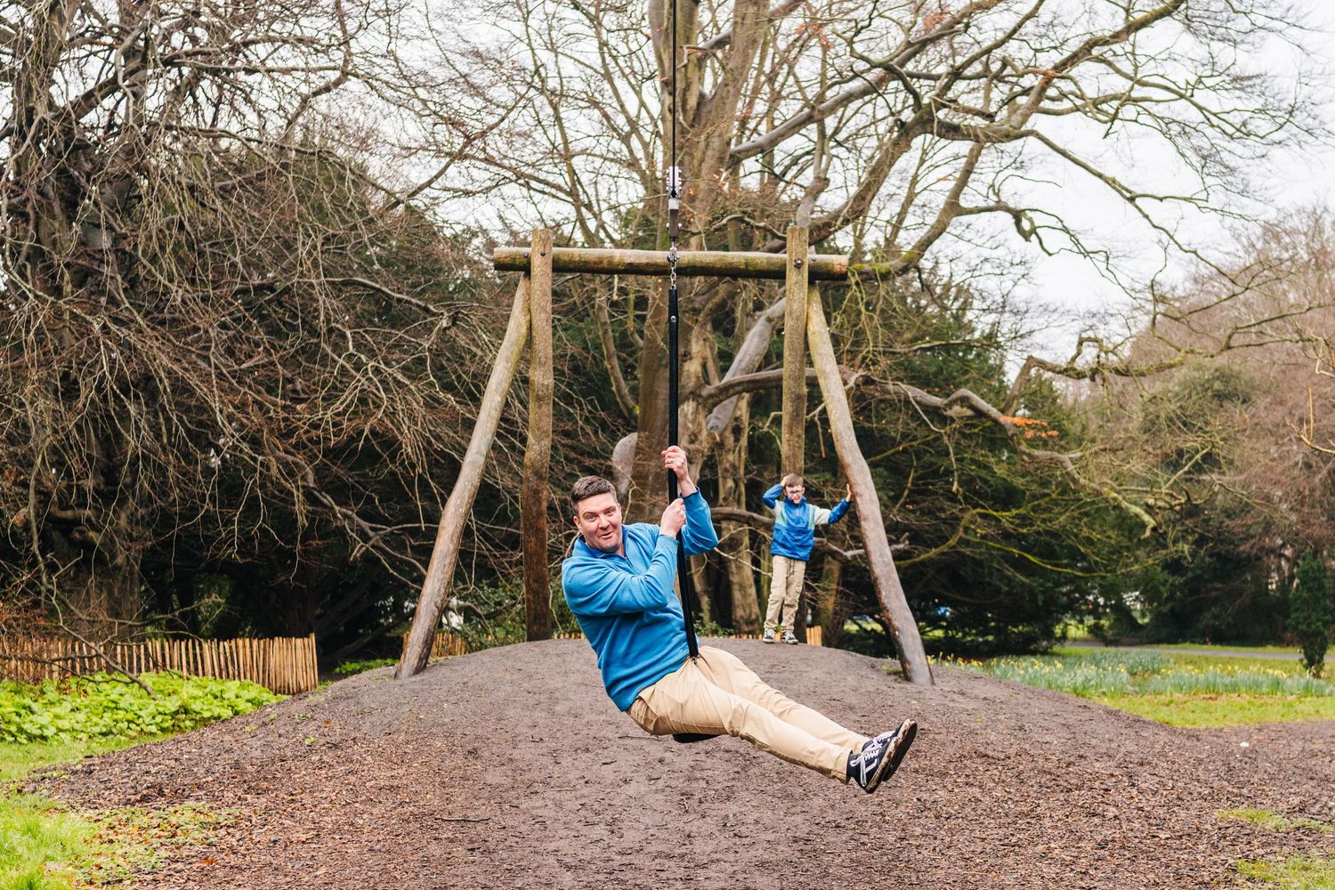 Family photographer Dublin A person wearing a blue jacket swings on a wooden swing set in an outdoor park setting with bare trees in winter.