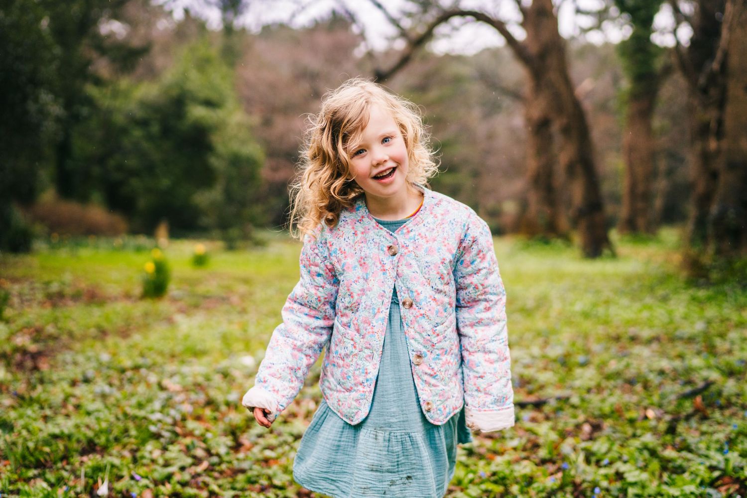 Child wearing floral quilted jacket and blue dress stands in a lush green park during autumn. Family photographer Dublin Ireland