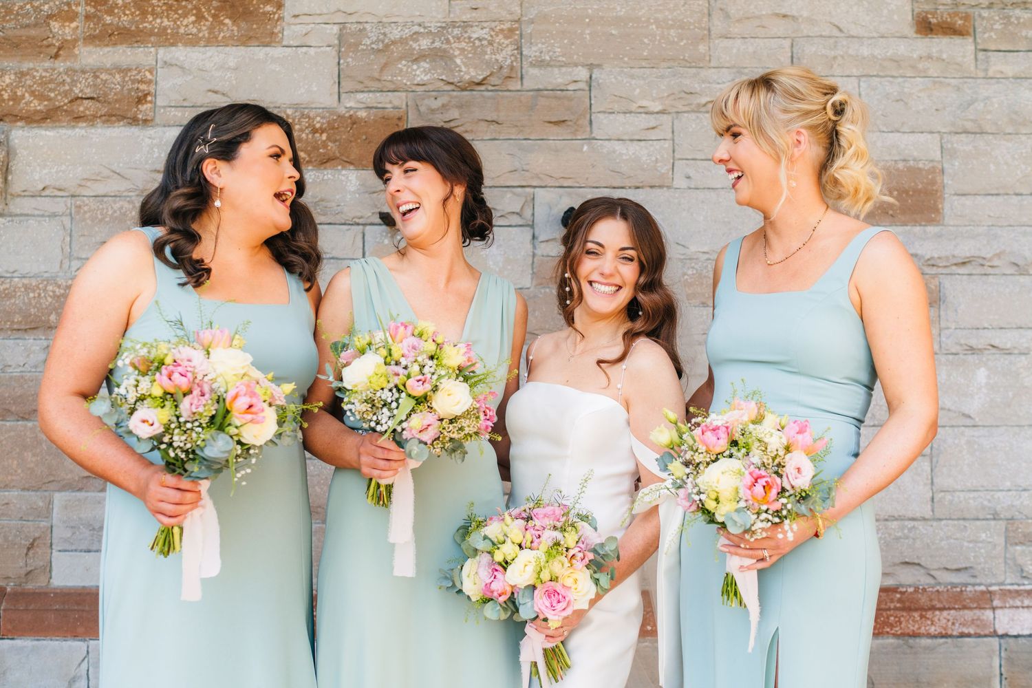 Four bridesmaids in soft mint green dresses share a candid laughing moment while holding pink and peach bouquets. Castle Leslie Wedding Phot