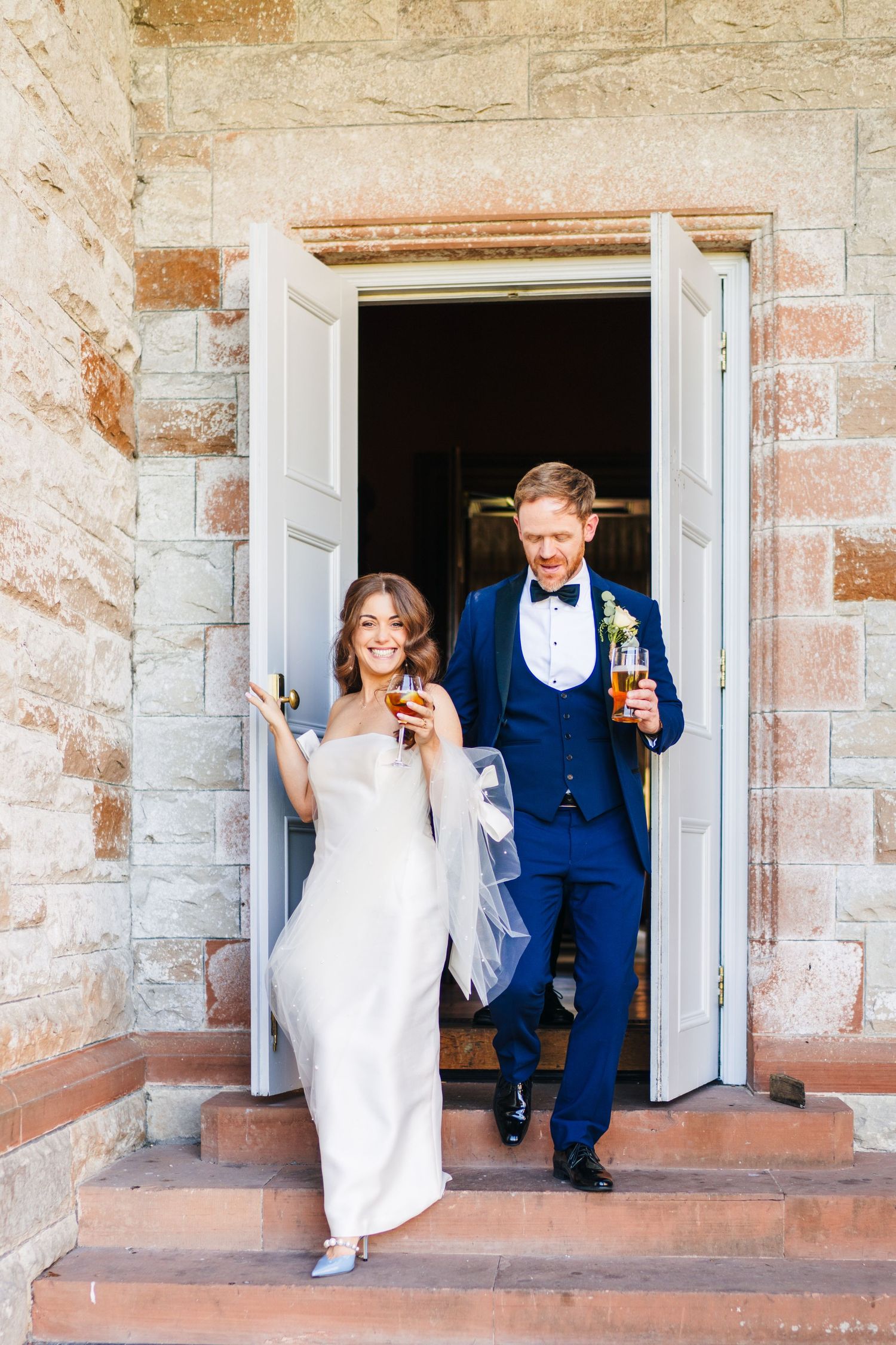Castle Leslie Wedding Photographer Dublin A wedding couple exits through white doors of a stone building, holding drinks and smiling.