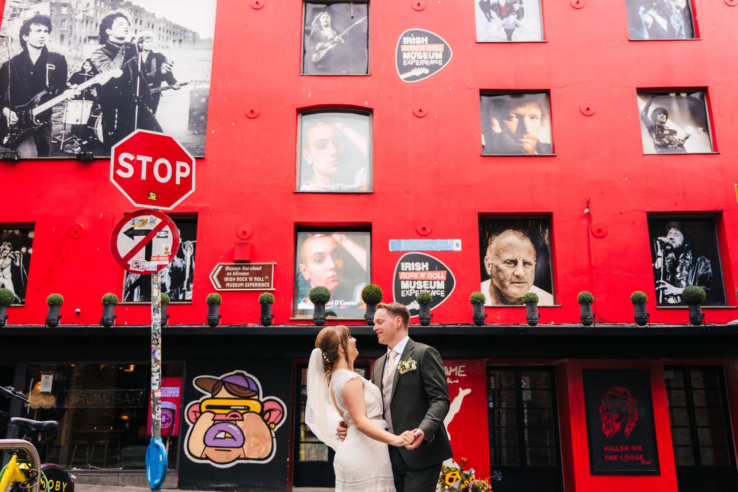 Wedding party poses in front of a bright red wall decorated with framed photographs. PHOTOGRAPHER Dublin Ireland Wedding