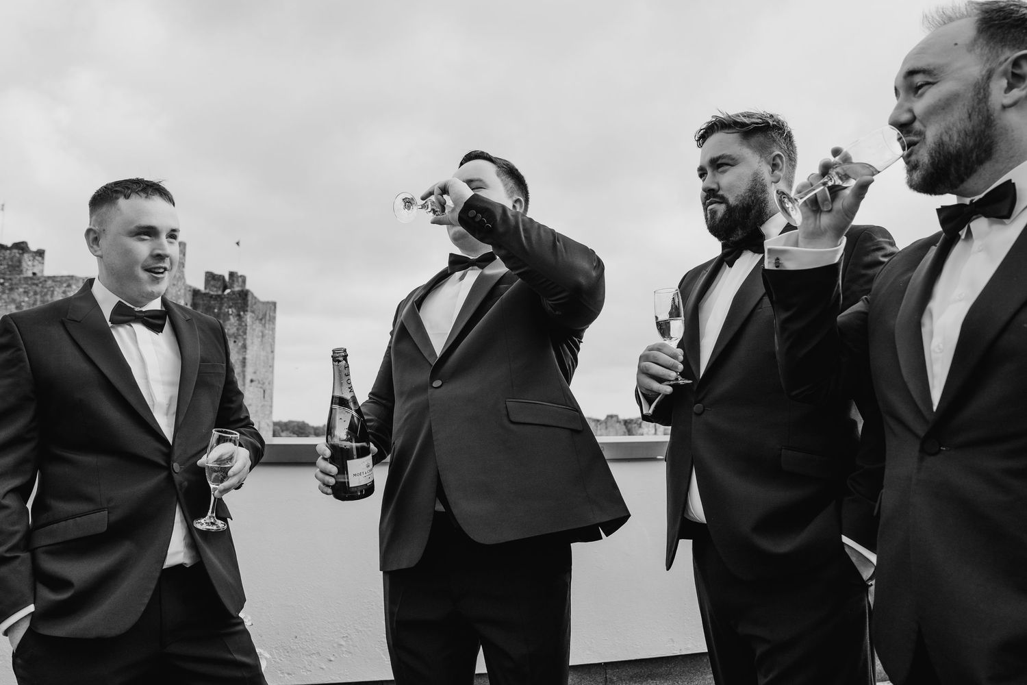 A group of groomsmen in formal tuxedos share drinks and laughs outdoors during a wedding celebration. Wedding Photography Trim Castle Irelan