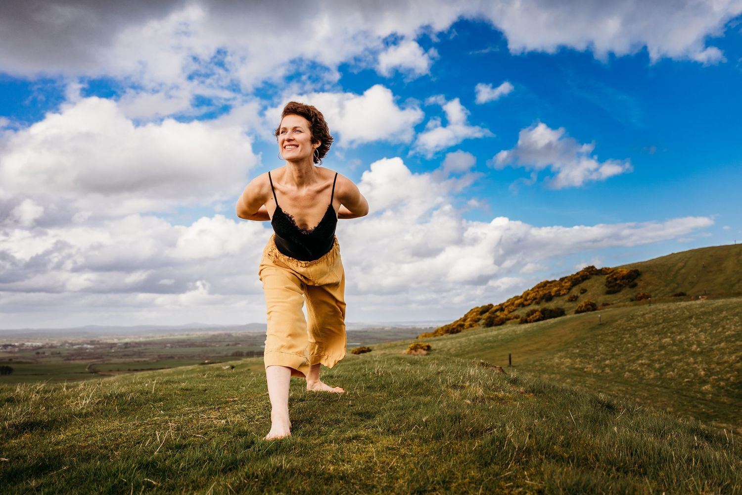 A person in a yellow dress runs joyfully across a green hillside under a dramatic blue sky with white puffy clouds. Portrait PHOTOGRAPHER Du