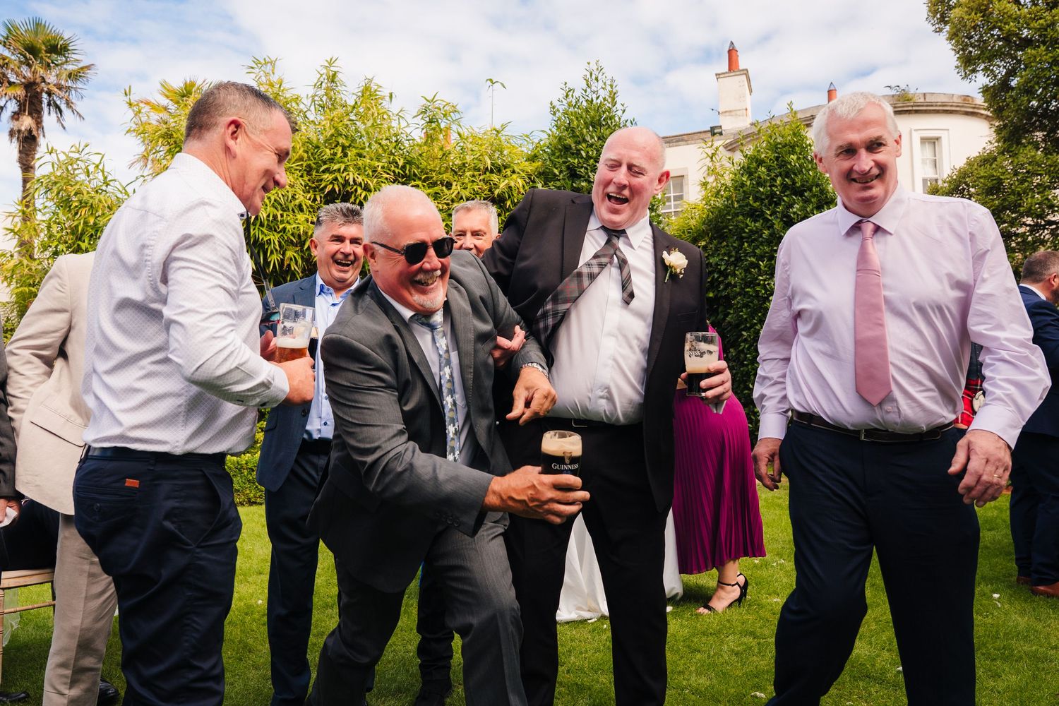 Groomsmen in formal attire pose playfully in a line outdoors on a sunny day. Portmarnock Hotel Wedding PHOTOGRAPHER Dublin