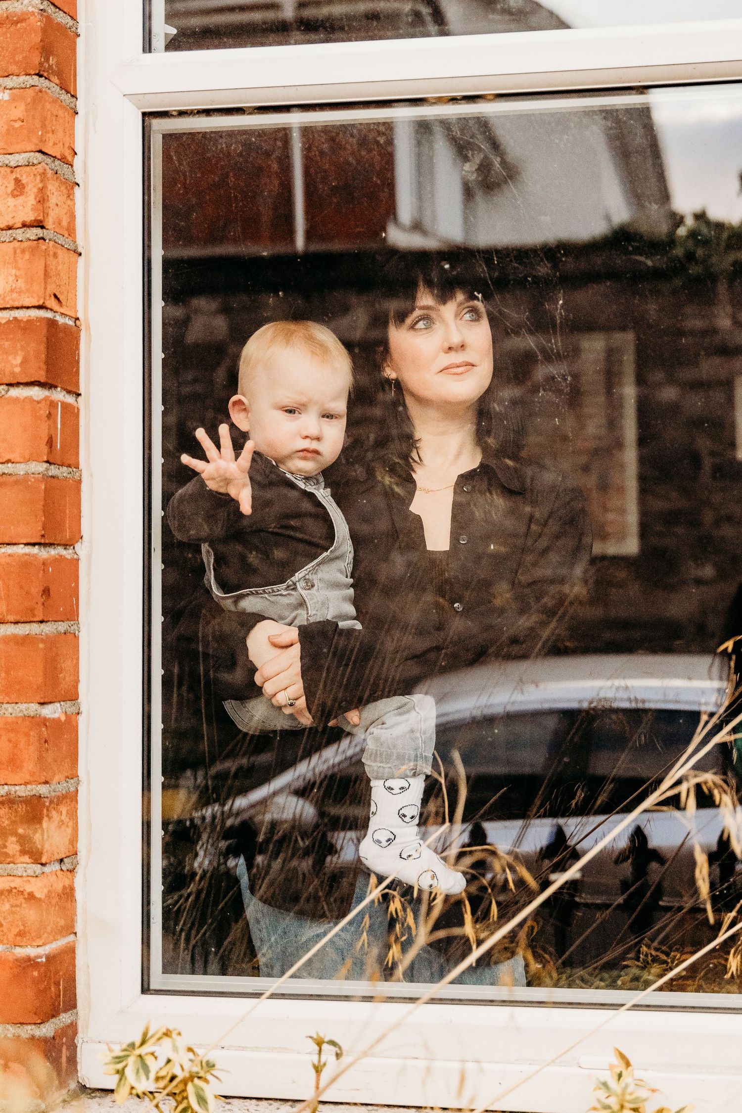 A tender moment captured through a window showing an adult holding a small child against a brick exterior backdrop.  Family PHOTOGRAPHER Dub
