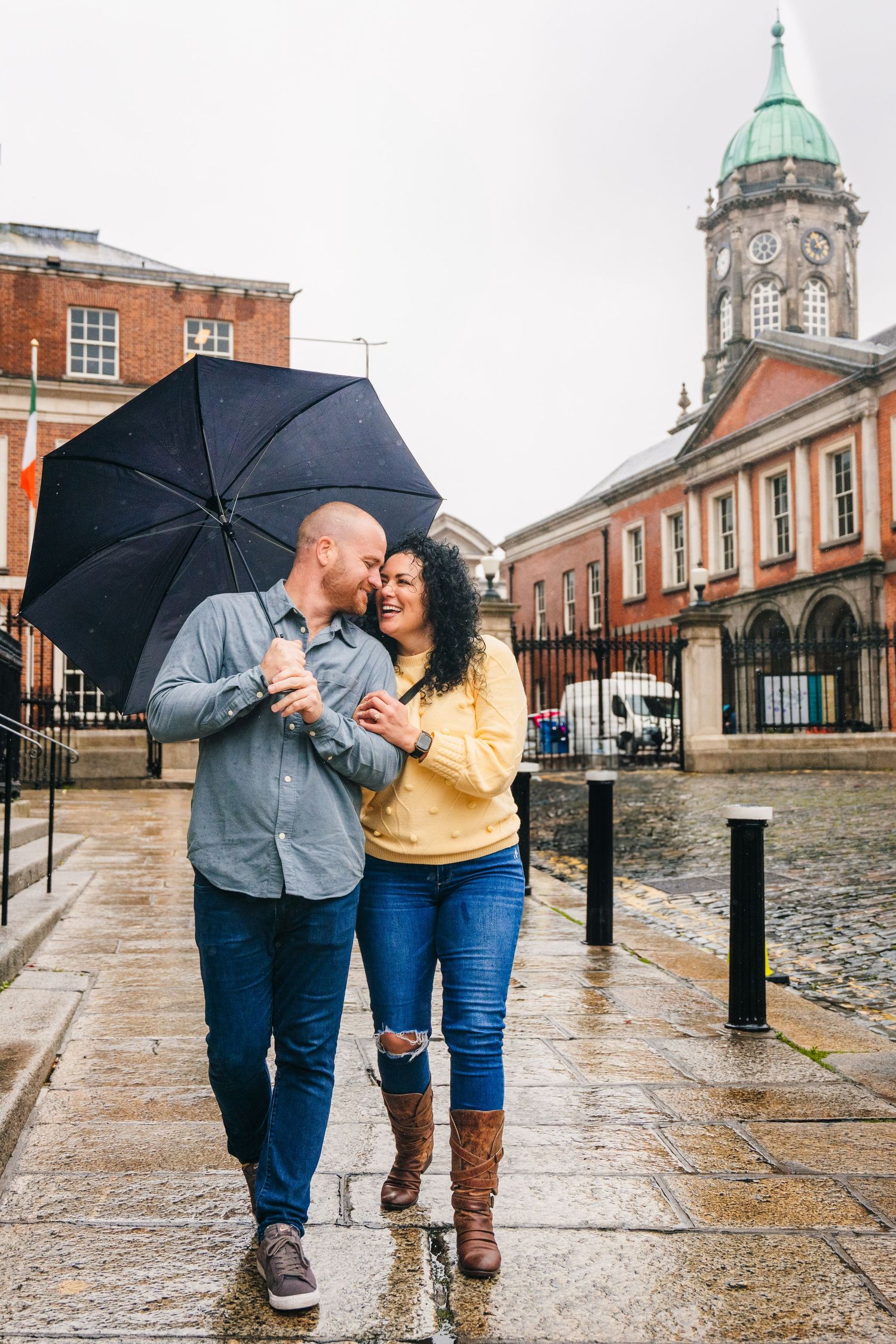 Couple laughing and walking on wet cobblestone street with black umbrella near historic architecture. Proposal Dublin Photographer