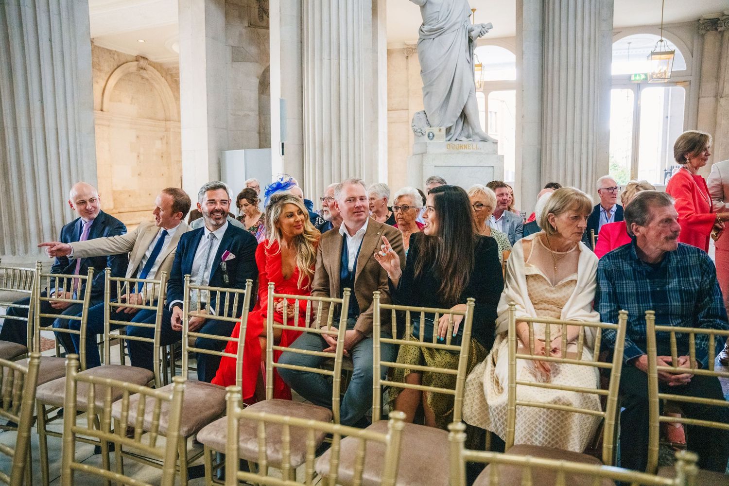 Wedding guests seated in elegant white chairs inside a grand columned venue with classical architecture.  city hall dublin wedding photos