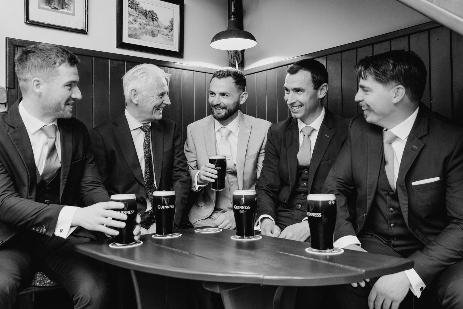 Five men in suits laughing and enjoying pints of Guinness together at a pub table in a black and white photo. Wedding photographer Dublin