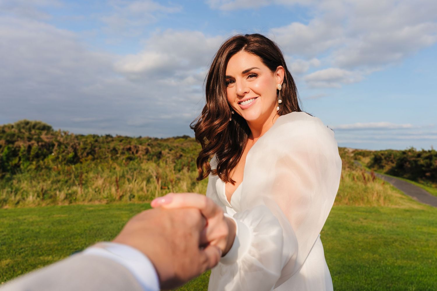 A couple in wedding attire embraces on a scenic hilltop path with green grass and cloudy skies in the Wedding Photographer dublin