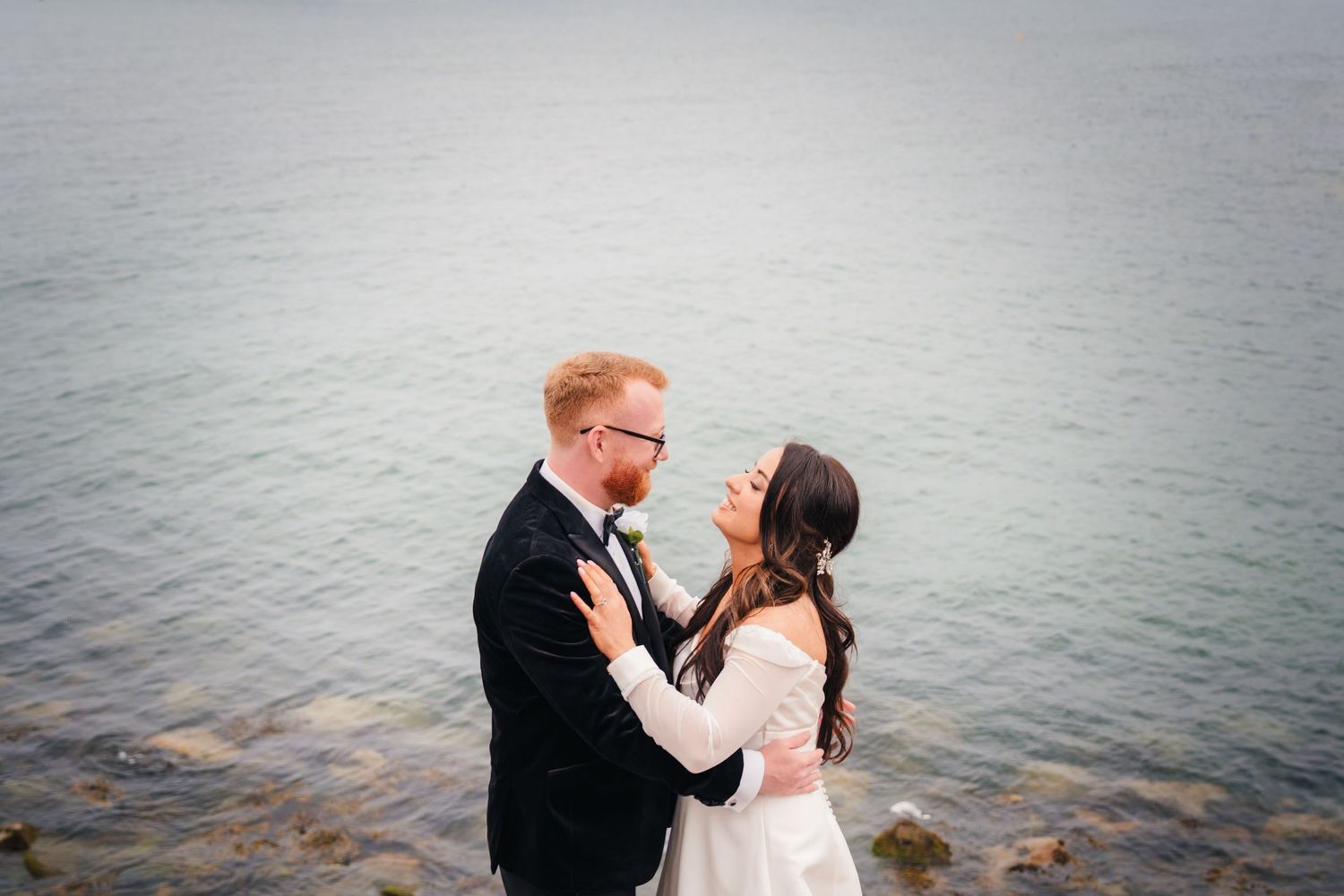 Bride and groom share a romantic moment by the ocean, with the groom in a suit and bride in a white gown.