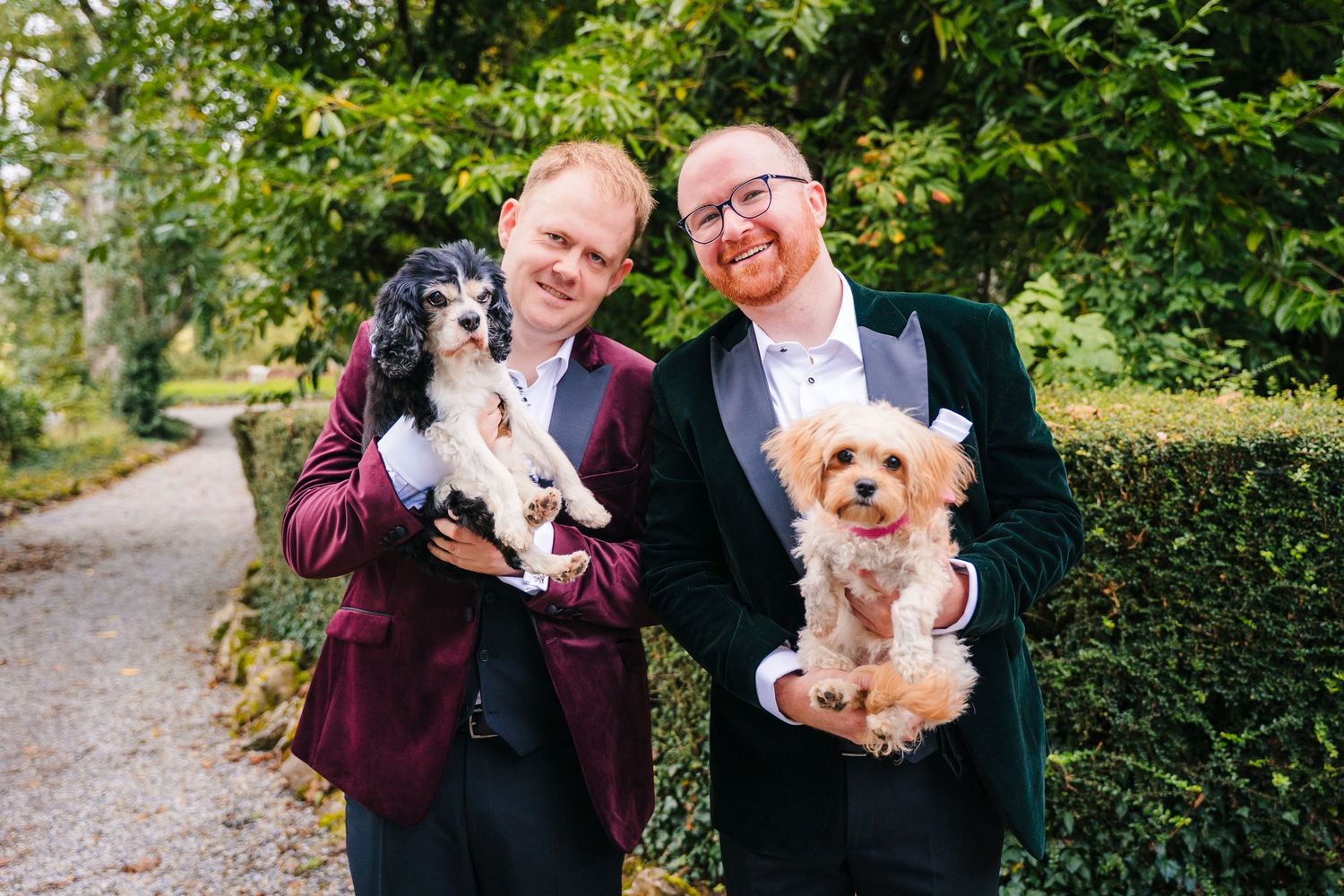 Two people in burgundy and black tuxedos holding small dogs on a garden path. LGBTQ Ashley Park Wedding Photographer